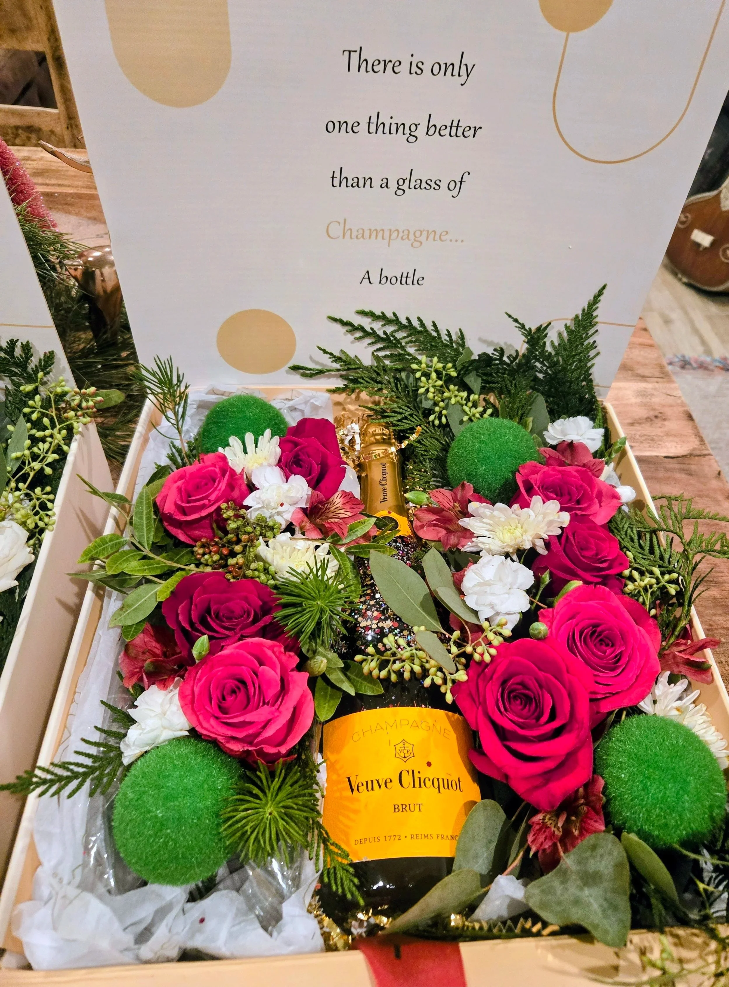 Gift box with a bottle of Veuve Clicquot champagne surrounded by pink roses, white flowers, green foliage, and green decorative balls, with a sign above that reads, 'There is only one thing better than a glass of Champagne... A bottle.'