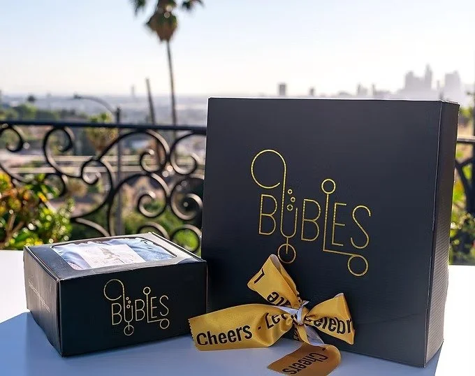 Celebration gift boxes labeled 'G Bubbles' with a yellow ribbon, set outdoors on a balcony with a city skyline and palm trees in the background.