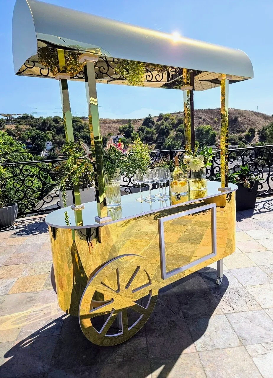 A gold-colored mobile bar cart with a gold canopy on a balcony, decorated with flowers and glassware, overlooking a scenic hillside.