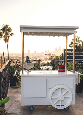 A white mobile cart with a canopy, wheels, and a large wheel design on the front, set up on an outdoor terrace with a city skyline in the background.