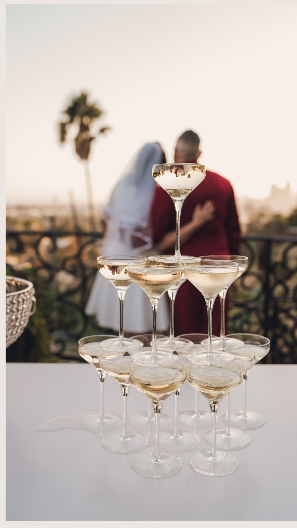 A pyramid of champagne glasses filled with champagne on a white table outdoors at sunset, with a blurred couple kissing in the background.
