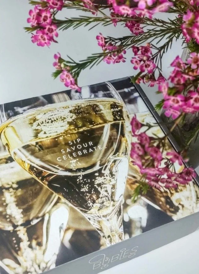 A close-up of a glass of champagne with the words 'Sip Savor Celebrate' written on it. There are pink flowers in the background.