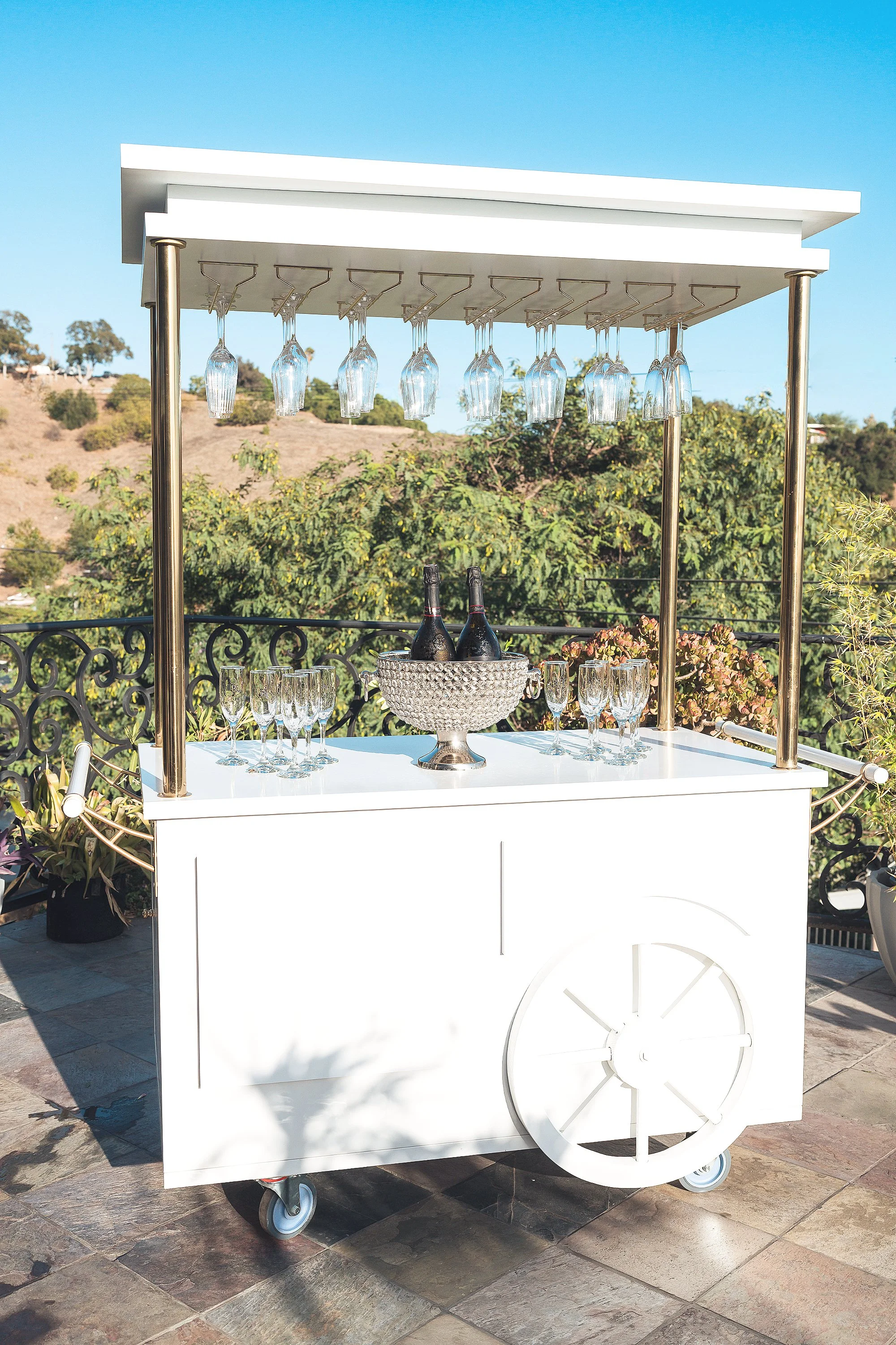 A white mobile bar cart with a canopy, featuring hanging glassware, bottles of champagne, and champagne glasses on top, set outdoors on a patio with a scenic landscape of trees, hills, and a clear blue sky in the background.