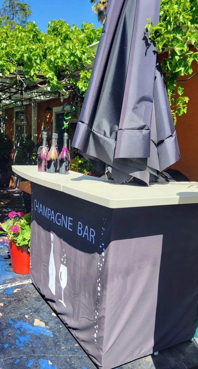 Outdoor champagne bar with bottles covered in pink glitter and an umbrella, surrounded by greenery and flowers.