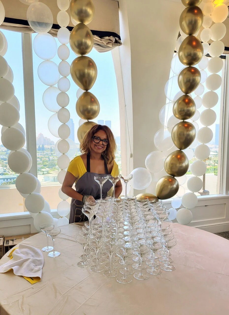 A woman with curly hair, glasses, and a yellow shirt, stands behind a table with a pyramid of empty champagne glasses, smiling, during a celebration or event in a room decorated with white and gold balloons.