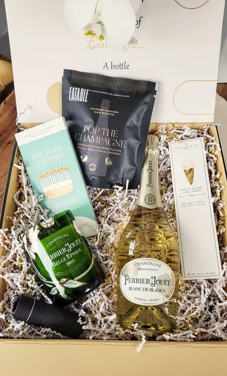 A gift box containing a bottle of Perrier-Jouet Blanc de Blancs champagne, a green candle with white flowers, a pack of gourmet popcorn labeled 'Pop the Champagne,' vanilla sugar cone crisps, and a birthday card.