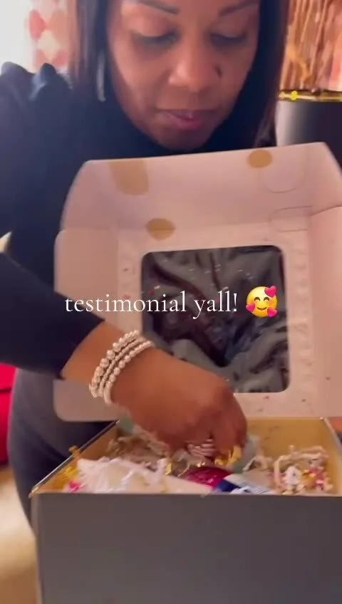 A woman with dark hair and wearing a black top, leaning over a gift box with a transparent window, inside which there are clothing items. She is pointing at the box, and the image has a caption that reads 'testimonial yall!' with a smiling face with hearts emoji.