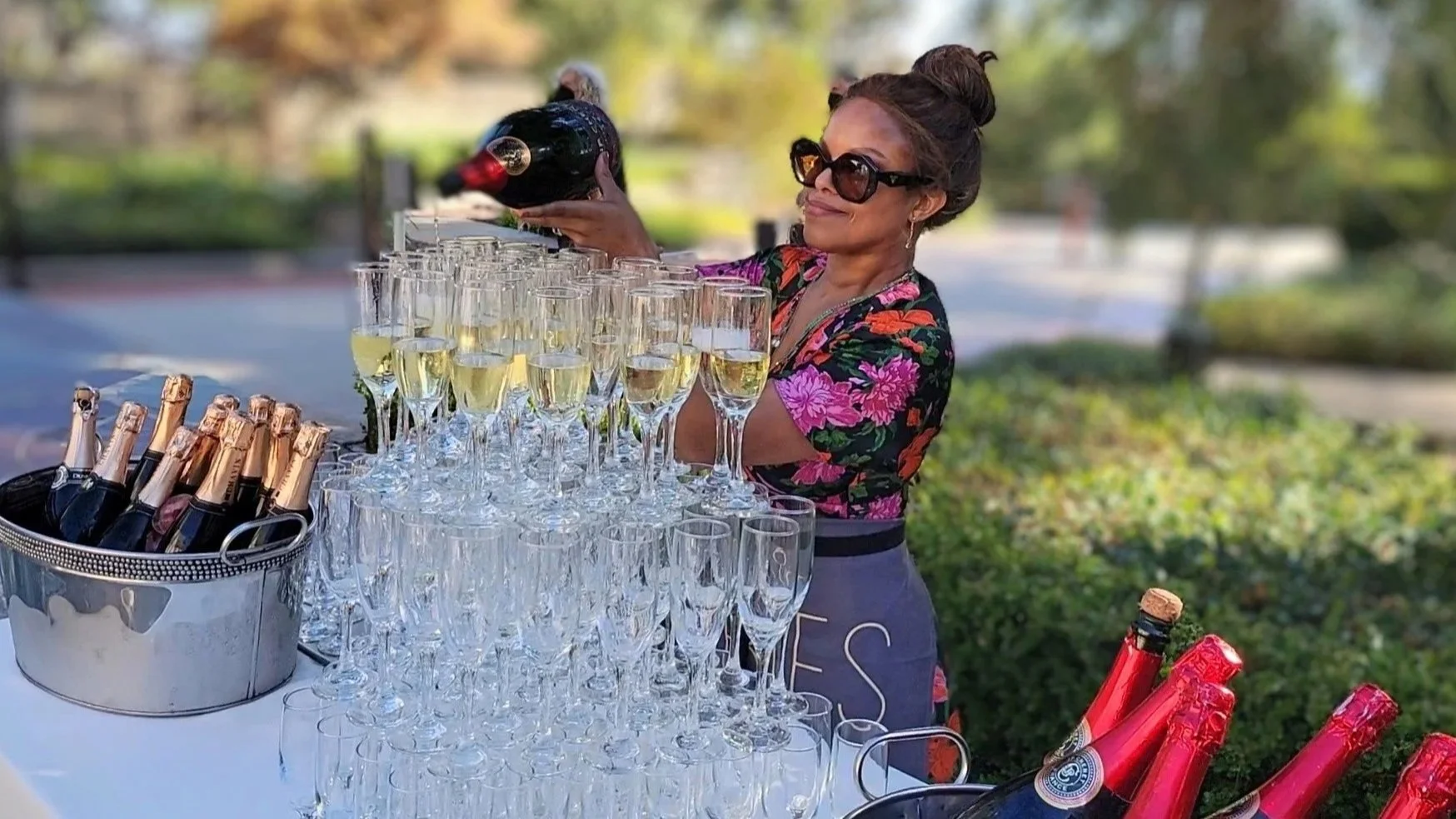 A woman in sunglasses pouring champagne into glasses at an outdoor party table with bottles of champagne and wine bottles nearby.