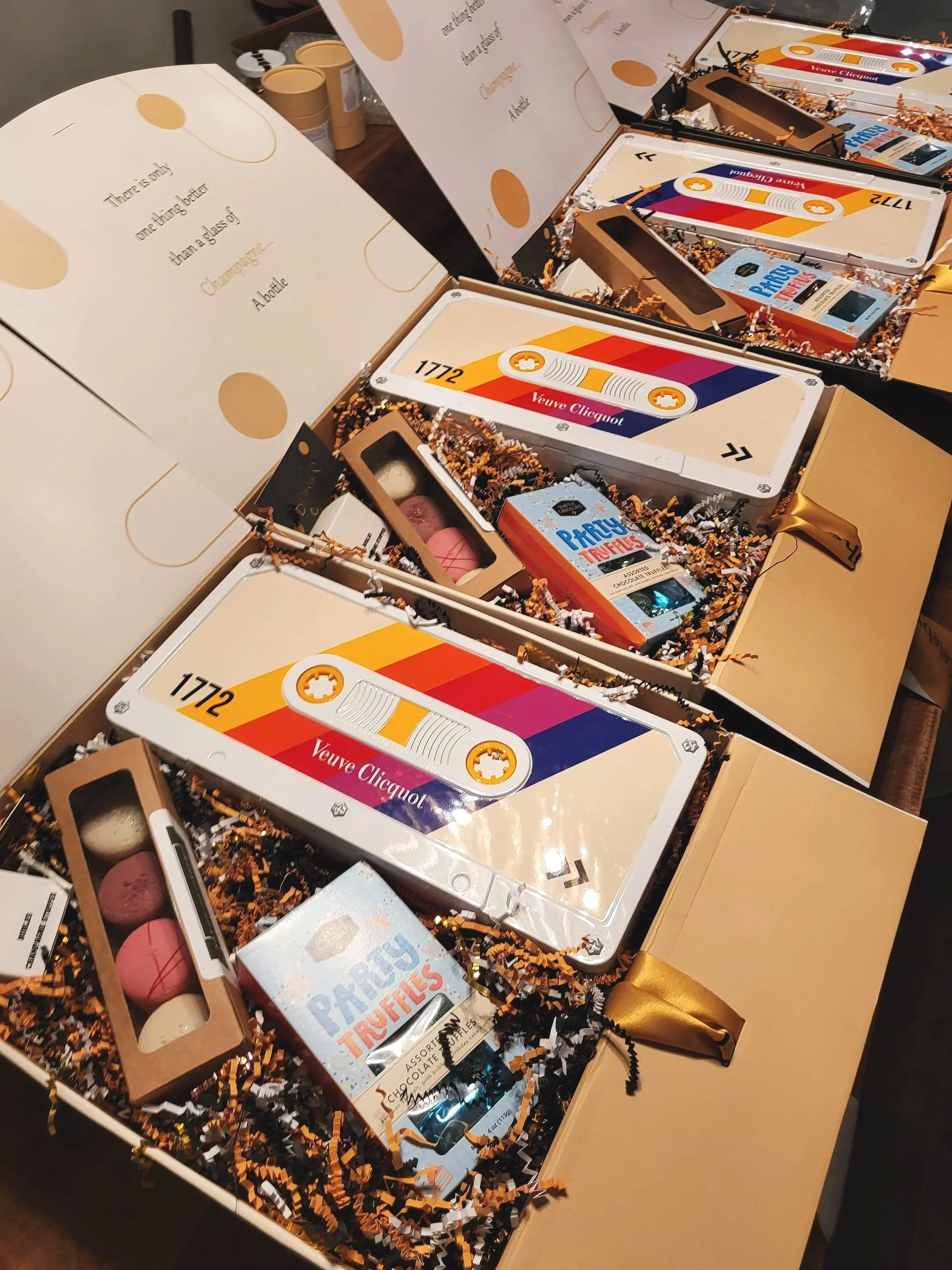 Open gift boxes with collectible items such as puzzle boxes, tins labeled Veuve Clicquot, and packages of Party Truffles chocolates, arranged on a table with shredded paper filler.