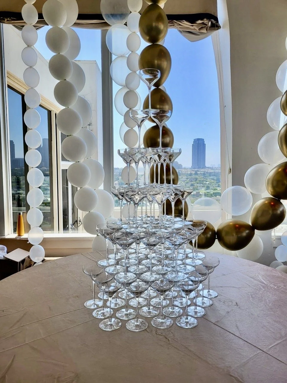 A tower of champagne glasses arranged in a pyramid shape, surrounded by white and gold balloons, with large windows and a cityscape view in the background.