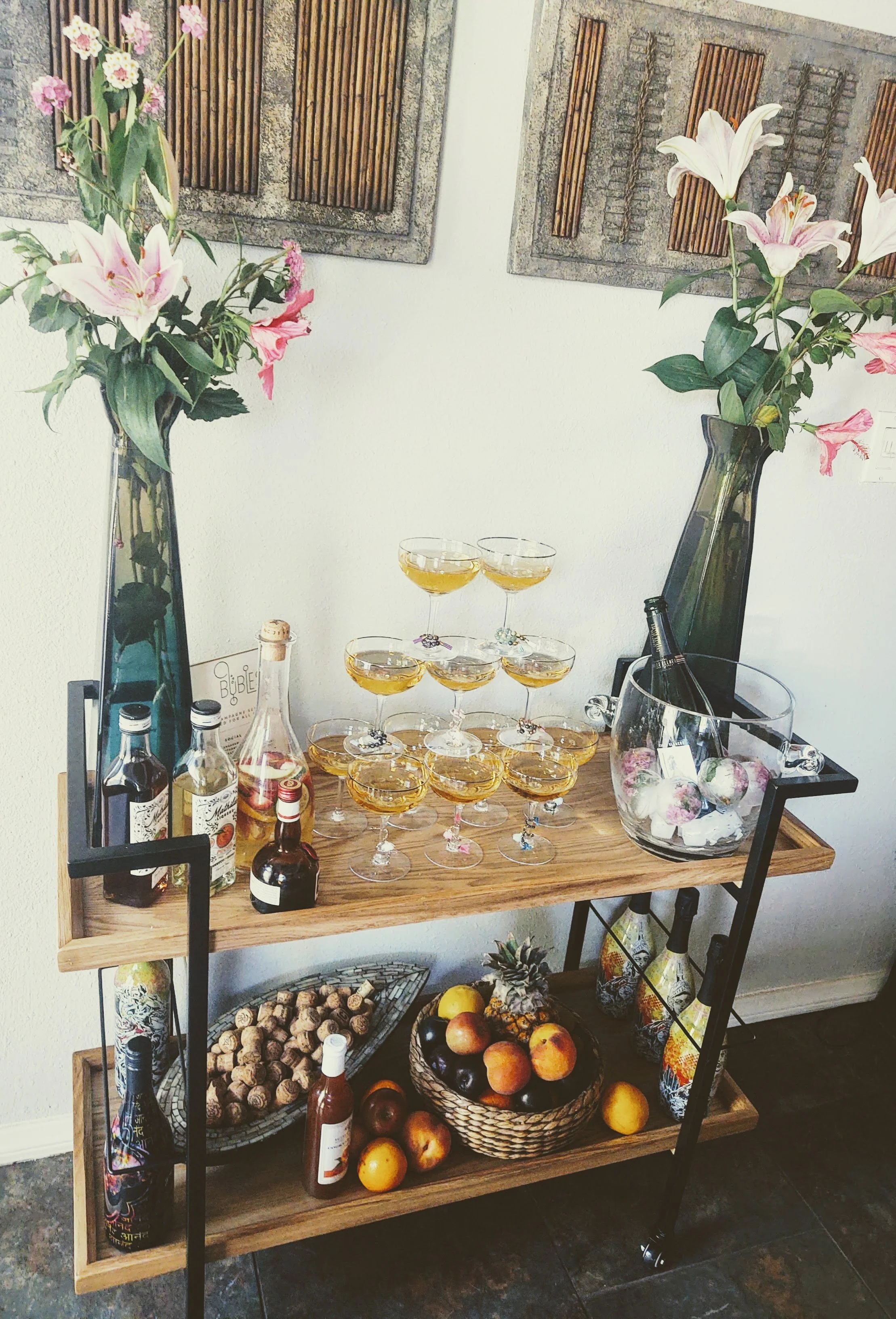 A wooden bar cart with bottles of alcohol, wine glasses filled with a yellow beverage arranged in a pyramid, and decorative items including flowers in tall vases, a bowl of apples and peaches, bottles of wine, and a basket of corks, against a wall decorated with two framed wooden art pieces with strips of wood in a pattern.
