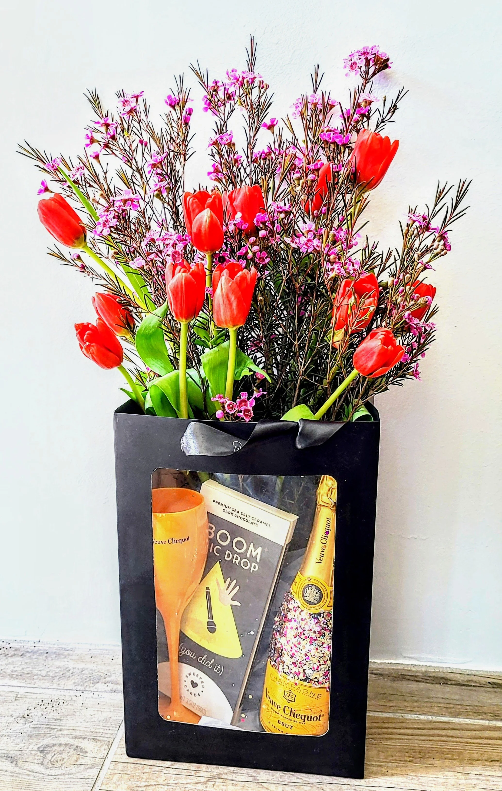 A black gift bag with a window showing a bottle of Veuve Clicquot champagne, a candle, and a card, containing a bouquet of red tulips and pink baby's breath flowers.