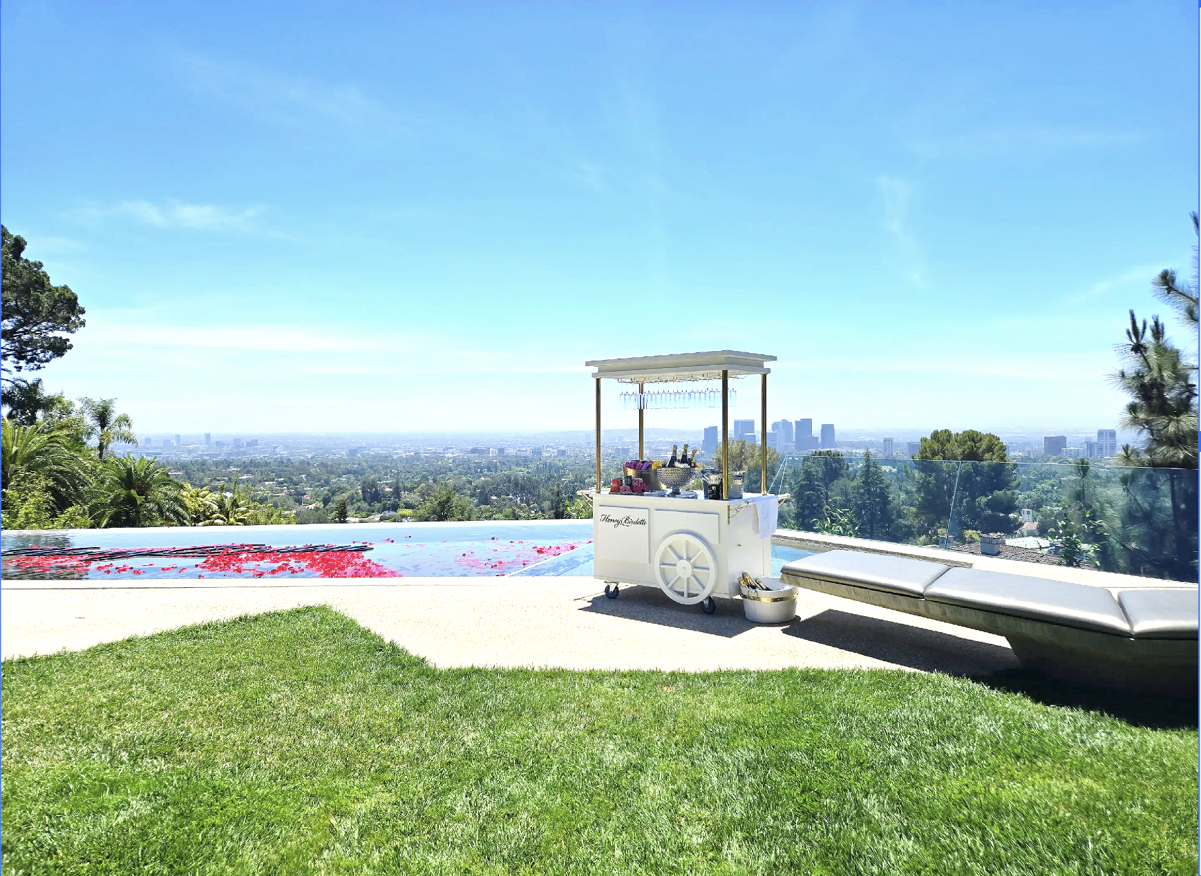A rooftop scene with a view of a city skyline in the distance, a Jacuzzi tub with rose petals, and a white mobile bar cart labeled "Honey Brushette" with drinks and accessories, alongside a lounge chair and green grass.
