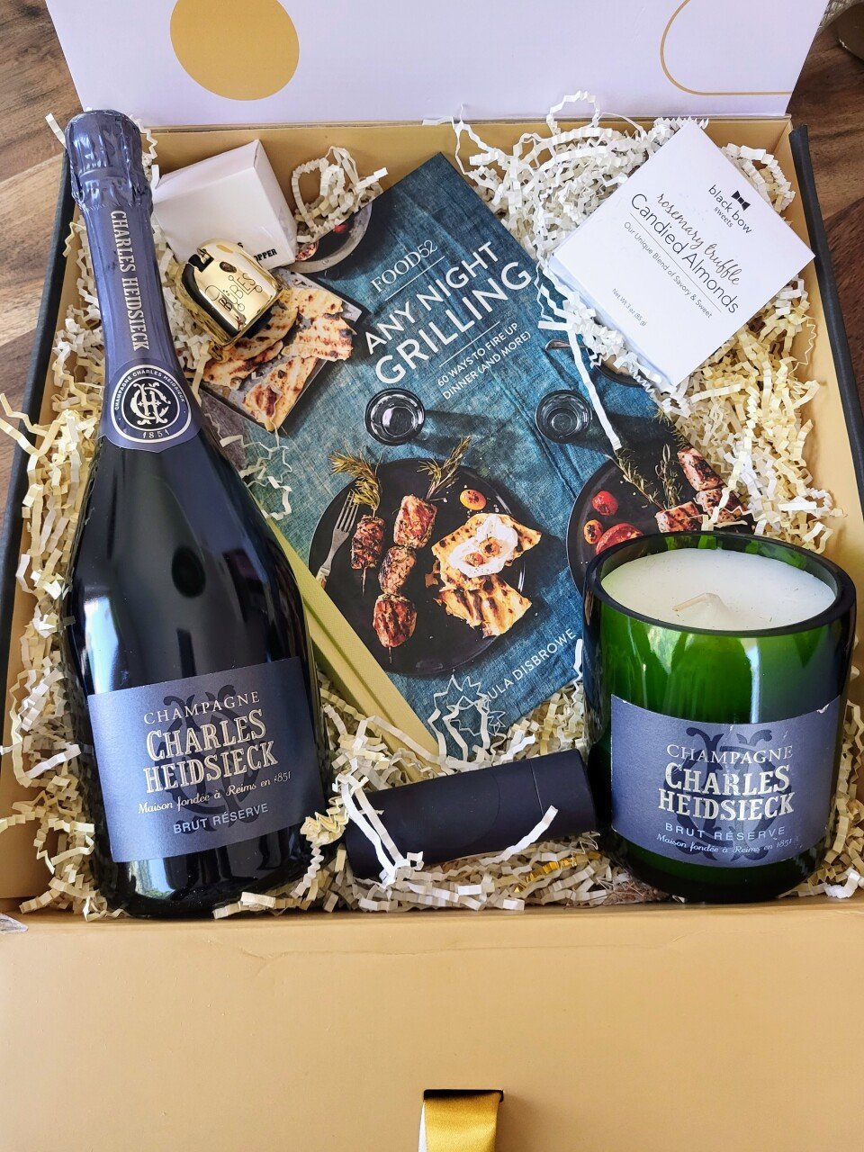 A gift box containing a bottle of Charles Heidsieck champagne, a green candle, a small wrapped item, a brochure titled 'Any Night Grilling,' a set of small soaps, a box of rosemary waffle cookies with almonds, and a paper with the logo 'black bowl' and some text about cookies and almonds, all nestled in shredded paper.
