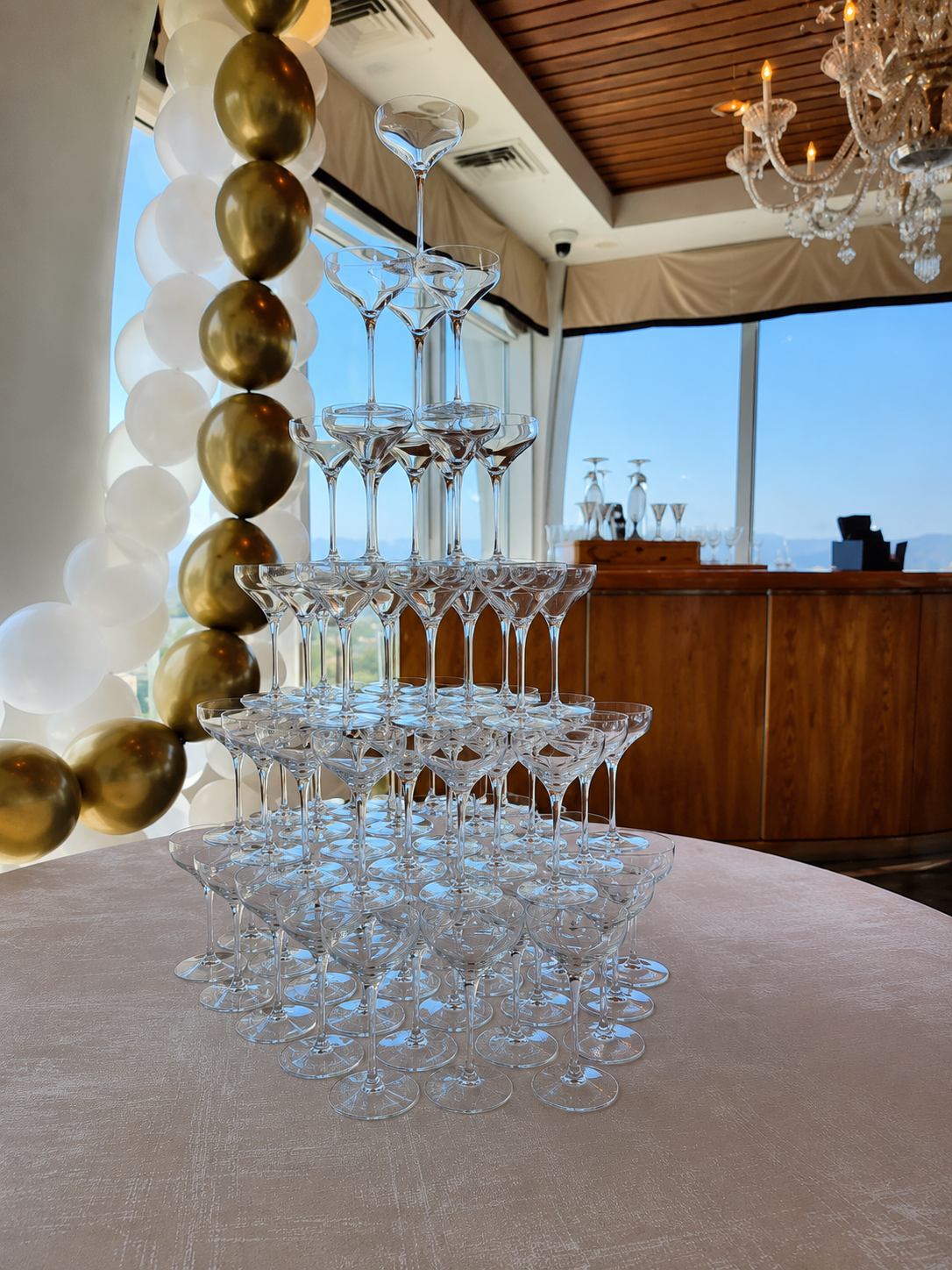 A pyramid of empty cocktail glasses arranged in a triangular formation on a table, with gold and white balloons and a chandelier in the background.