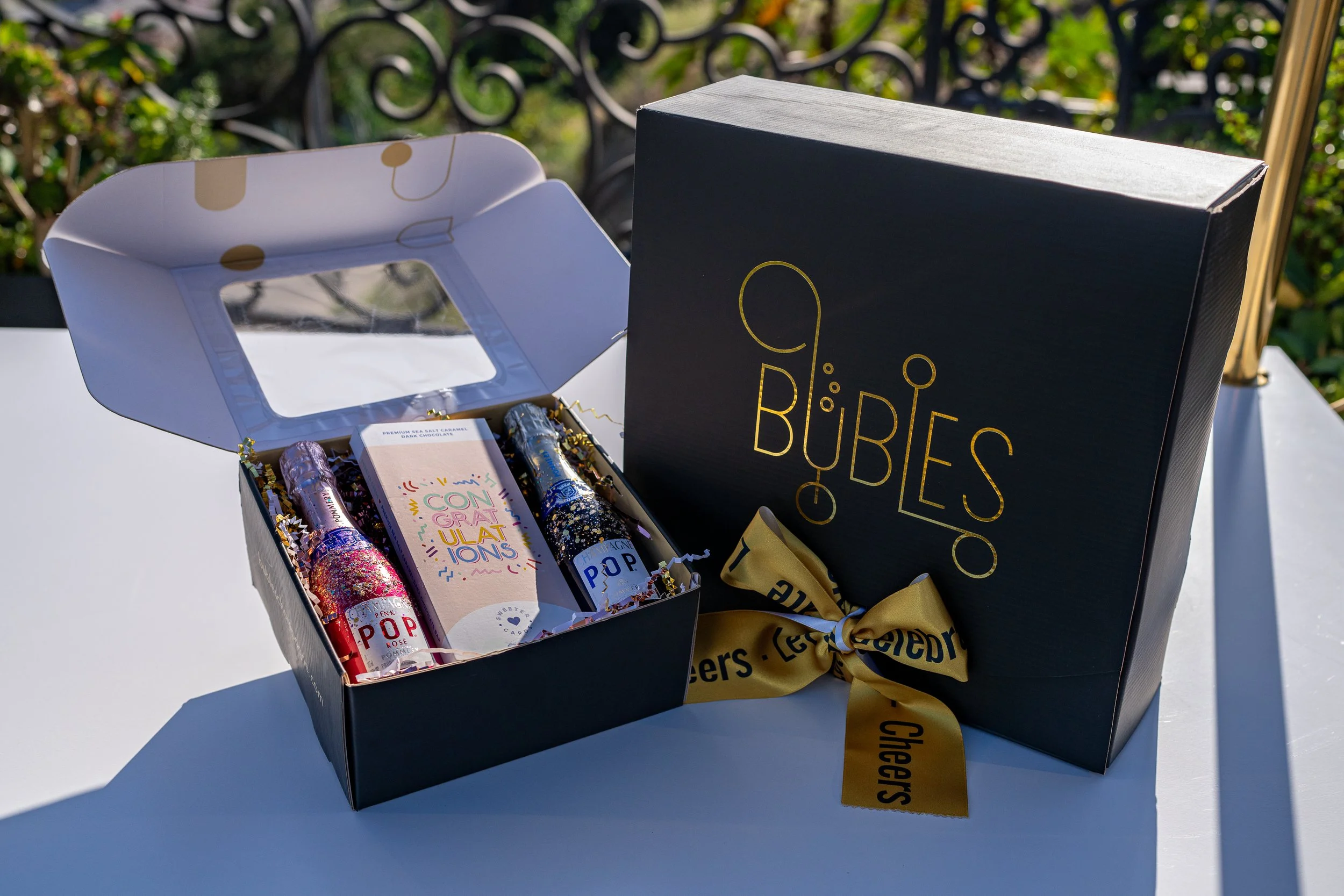 A black box gift set with a gold ribbon and a black box with gold lettering reading 'Chiques' is open to reveal two tubes of popcorn and other items with colorful packaging, set outdoors on a white surface.