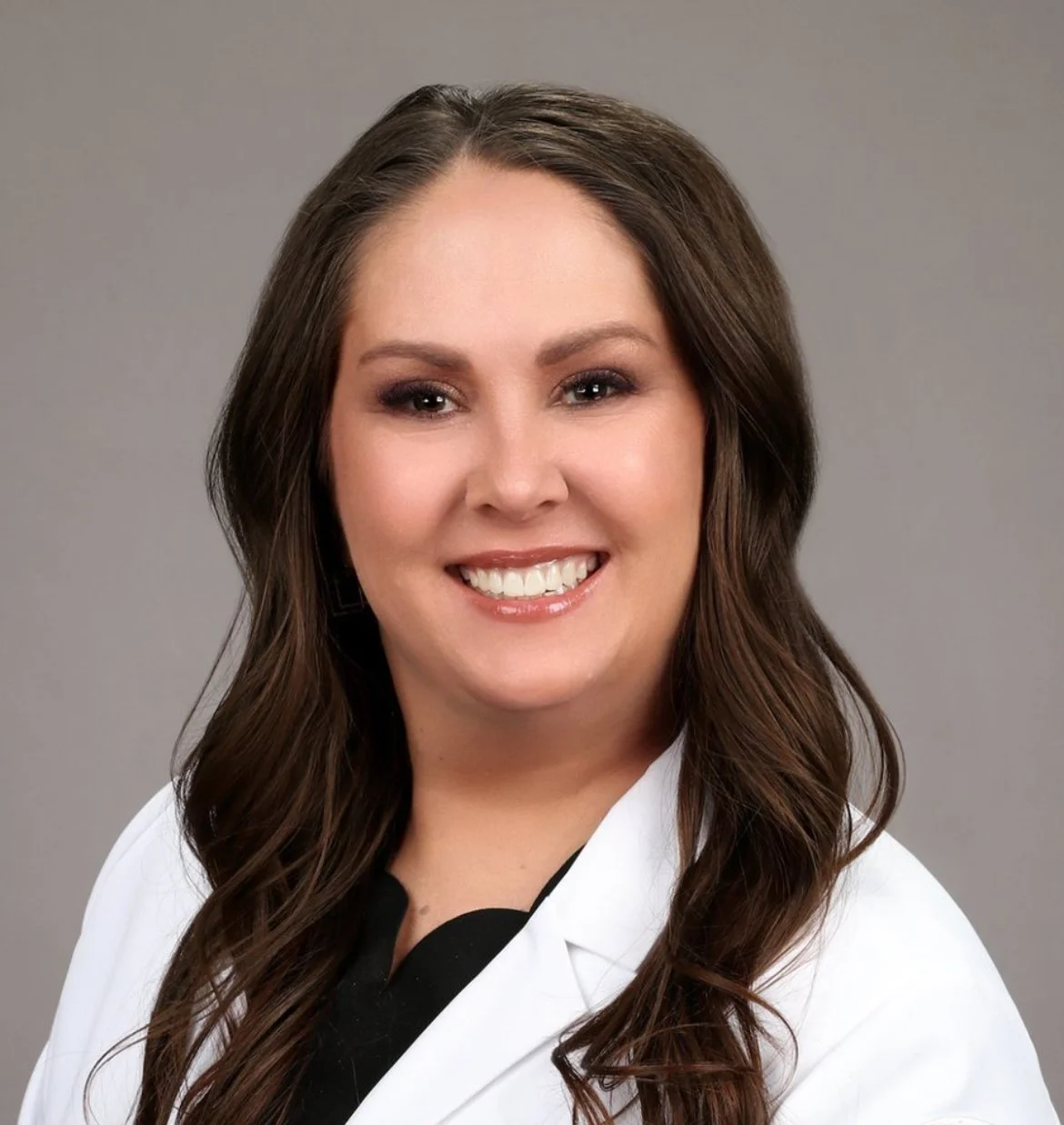 Professional woman with long brown hair smiling, wearing a white coat and black top against gray background.