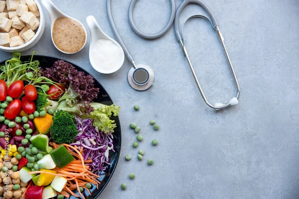 Fresh salad with vegetables and dressings, a stethoscope, and small scattered green peas on a gray surface.