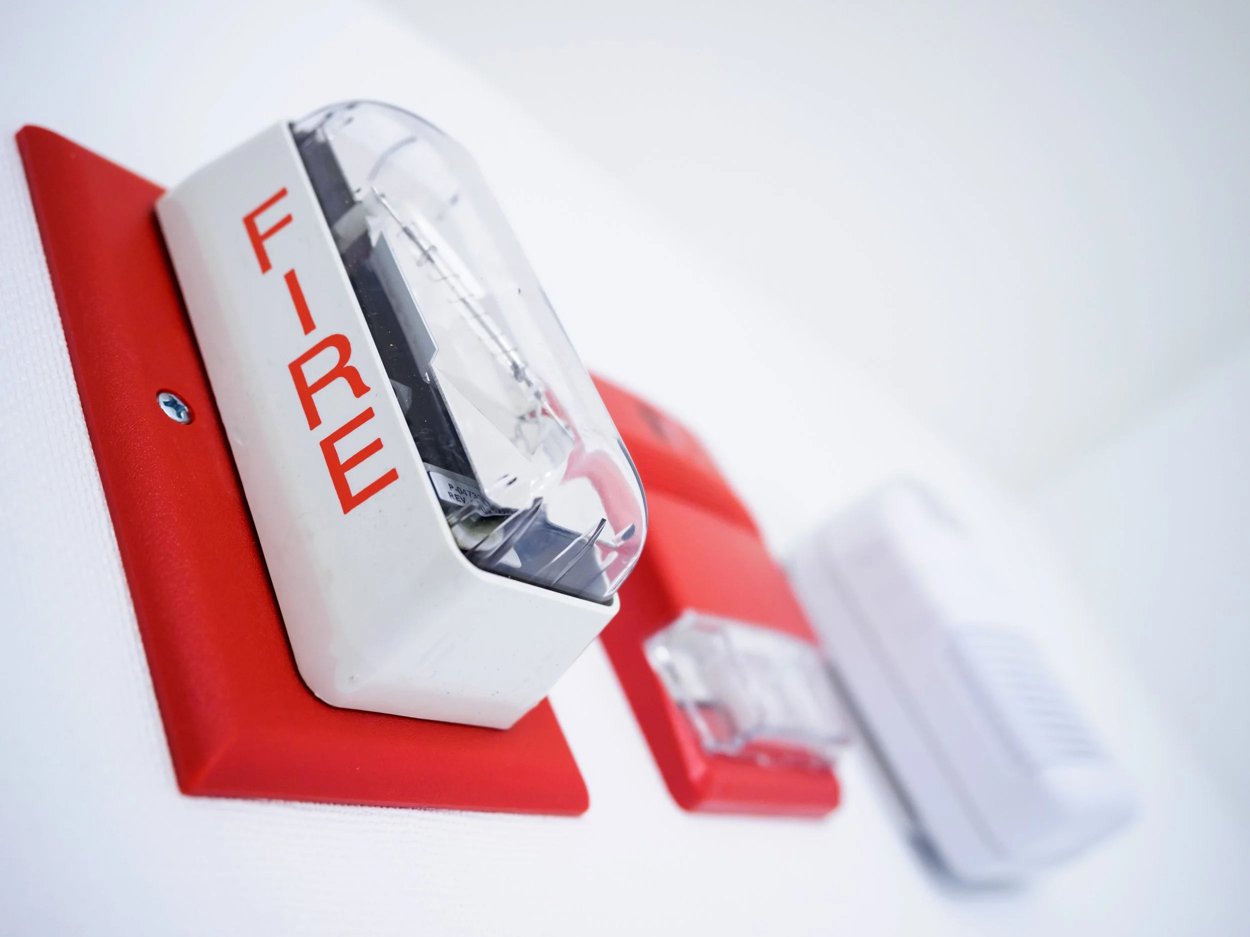 Close-up of a red and white fire alarm with a clear plastic cover and the word 'FIRE' written in red on the side, mounted on a white surface.