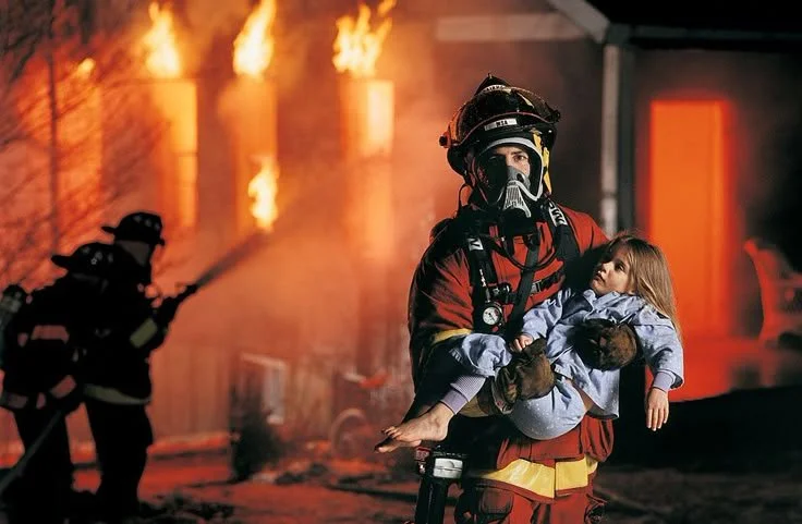 Firefighter carrying a young girl away from a blazing building fire at night.