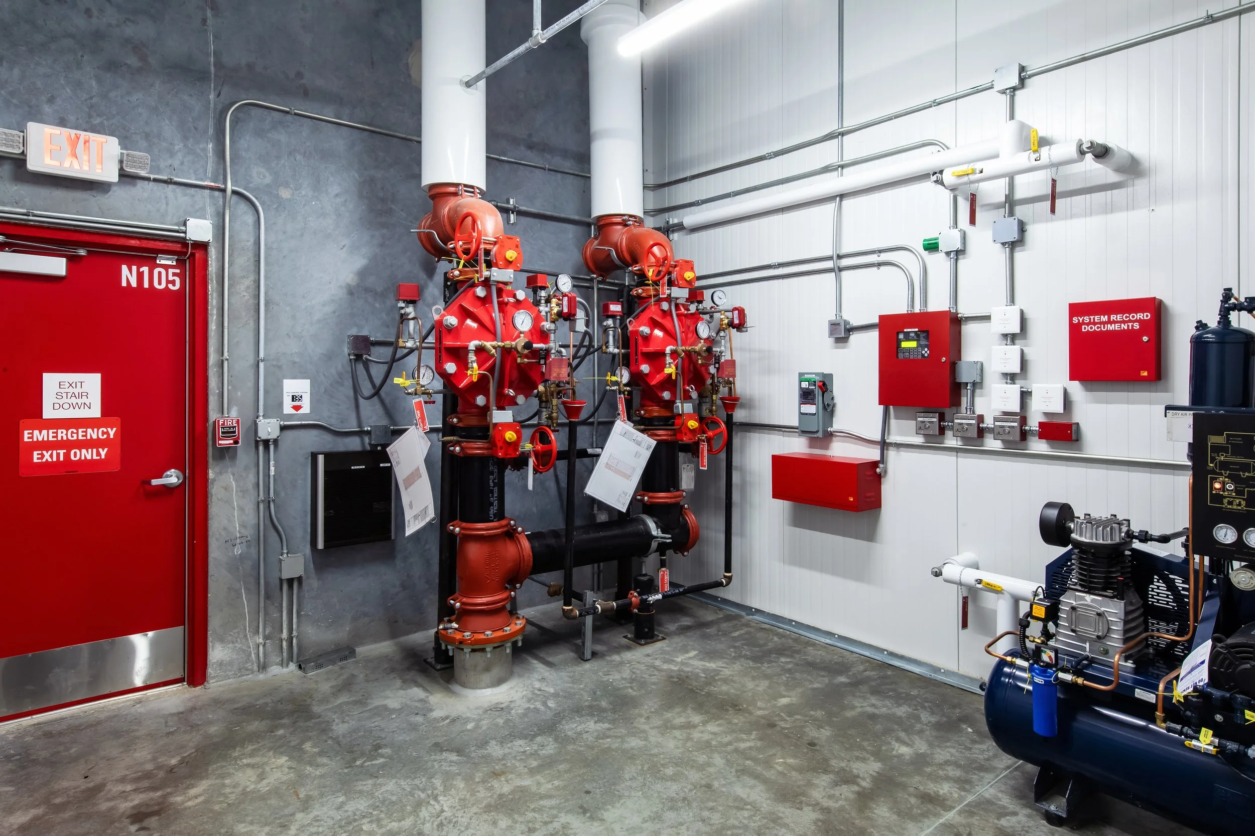 Industrial fire suppression system with red piping and gauges, emergency exit door, electrical boxes, and a compressor in a utility room.