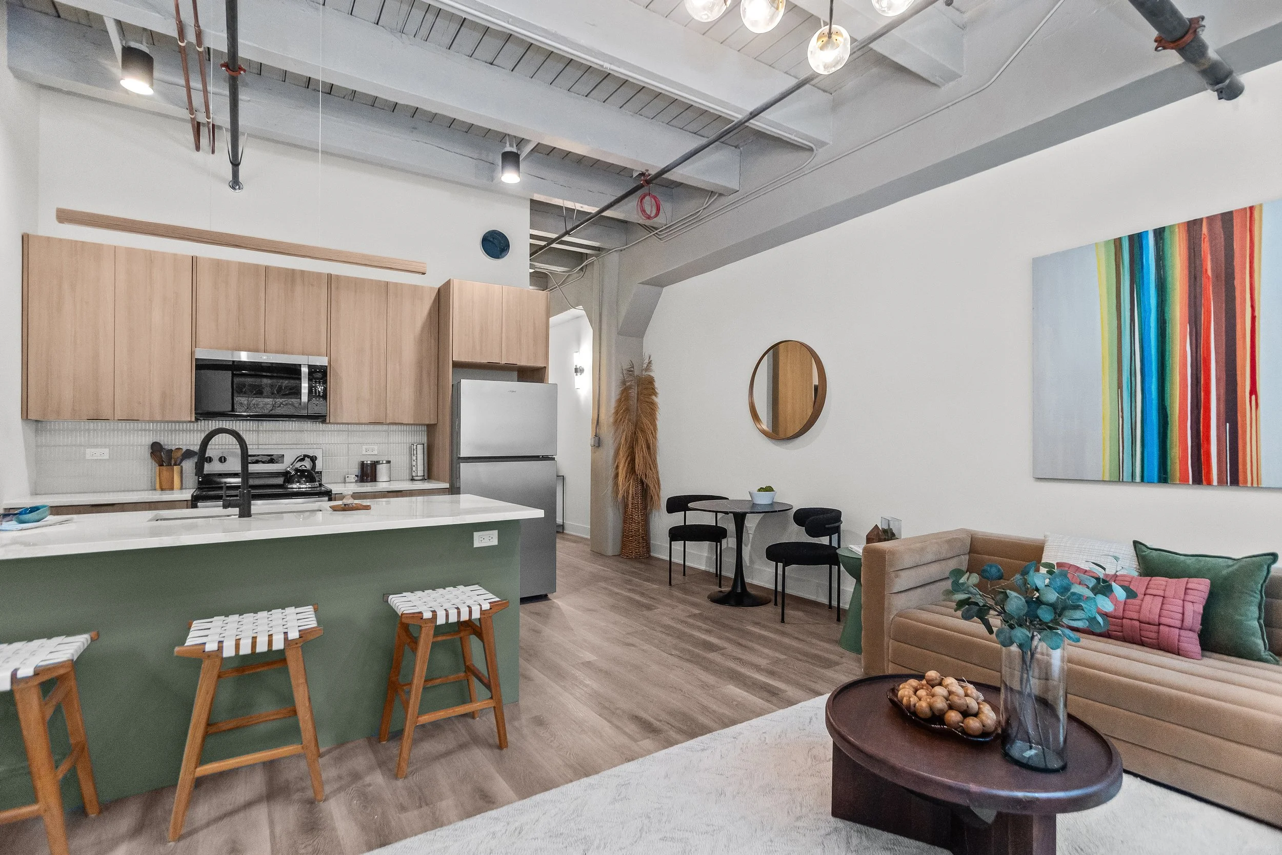 Open-concept living room and kitchen with modern decor, wooden cabinets, a green kitchen island with stools, a beige sofa with colorful pillows, a round coffee table with a vase and decorative objects, and a small dining area with a round table and three chairs. Artworks and a mirror adorn the white walls, and ceiling lights illuminate the space.