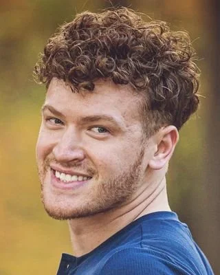 Young man with curly hair smiling outdoors in a blue shirt.