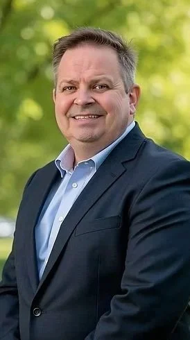 A man in a navy suit and light blue shirt standing outdoors with blurred green foliage in the background.