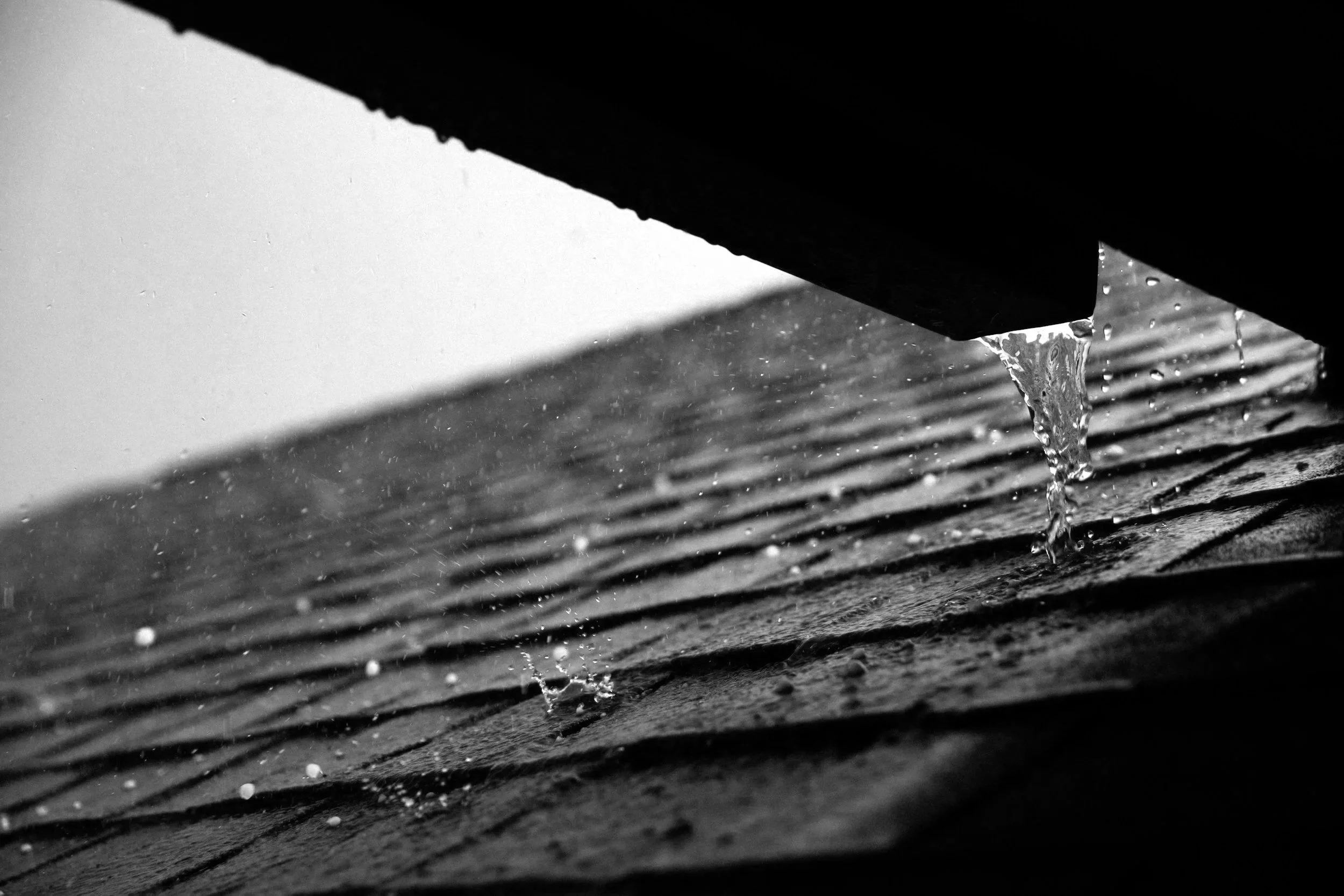 Close-up of a rainwater drop falling from the edge of a gutter onto a wooden surface with textured grooves.