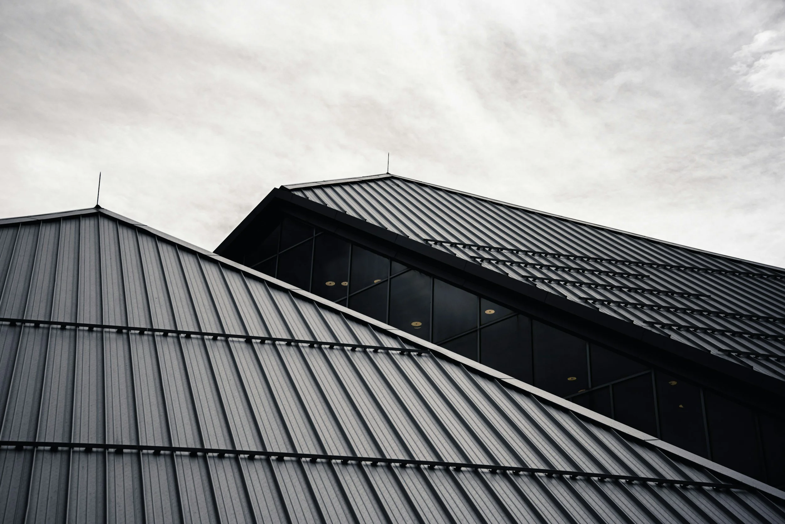 Close-up of a modern building with metal siding and large glass windows, topped with a dark sloped roof, against a cloudy sky.