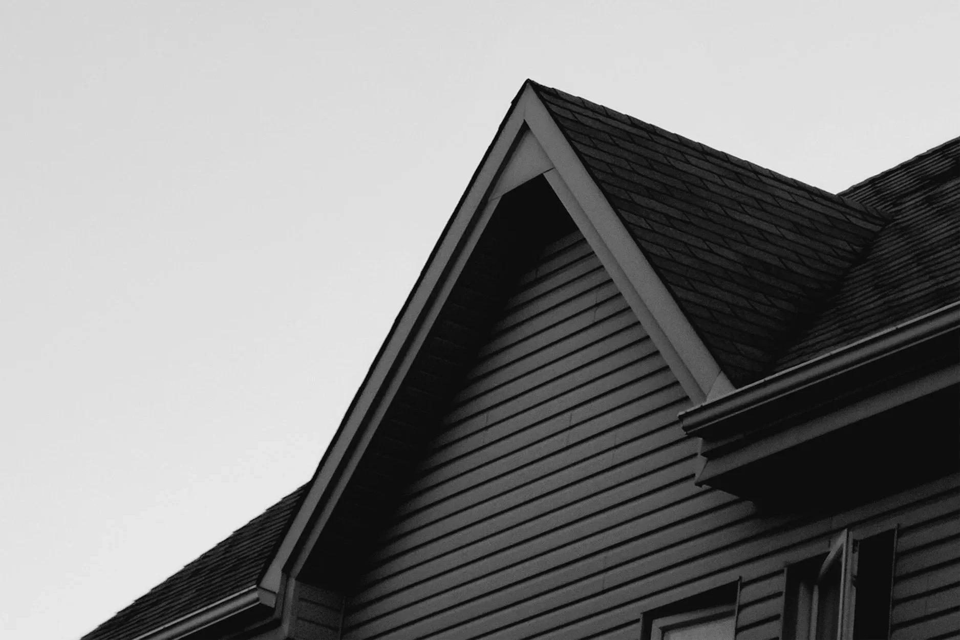 Close-up of the gable roof and siding of a house, with dark shingles and horizontal wooden exterior panels, against a plain, overcast sky.