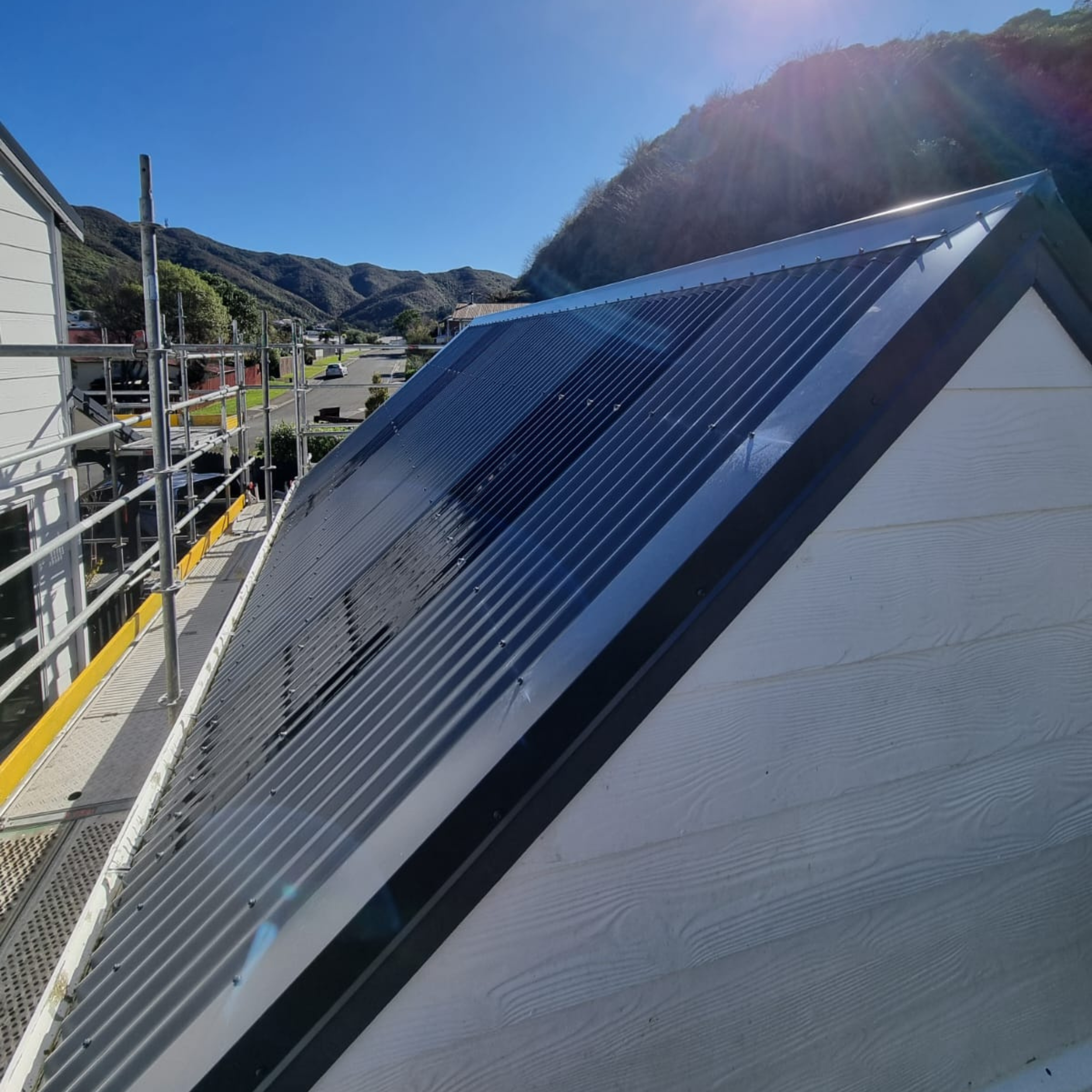 Roof of a building with metal panels and a scaffolding on the side, hillside with trees and small buildings in the background, clear blue sky, sunlight, and lens flare.