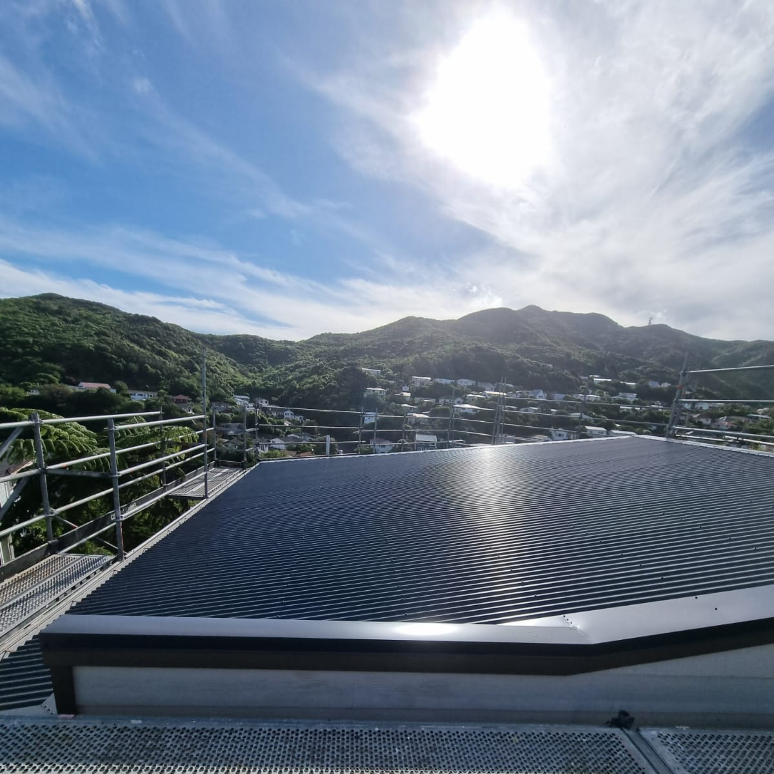 A metal balcony or walkway overlooking a landscape of green hills under a bright blue sky with some white clouds and the sun shining.