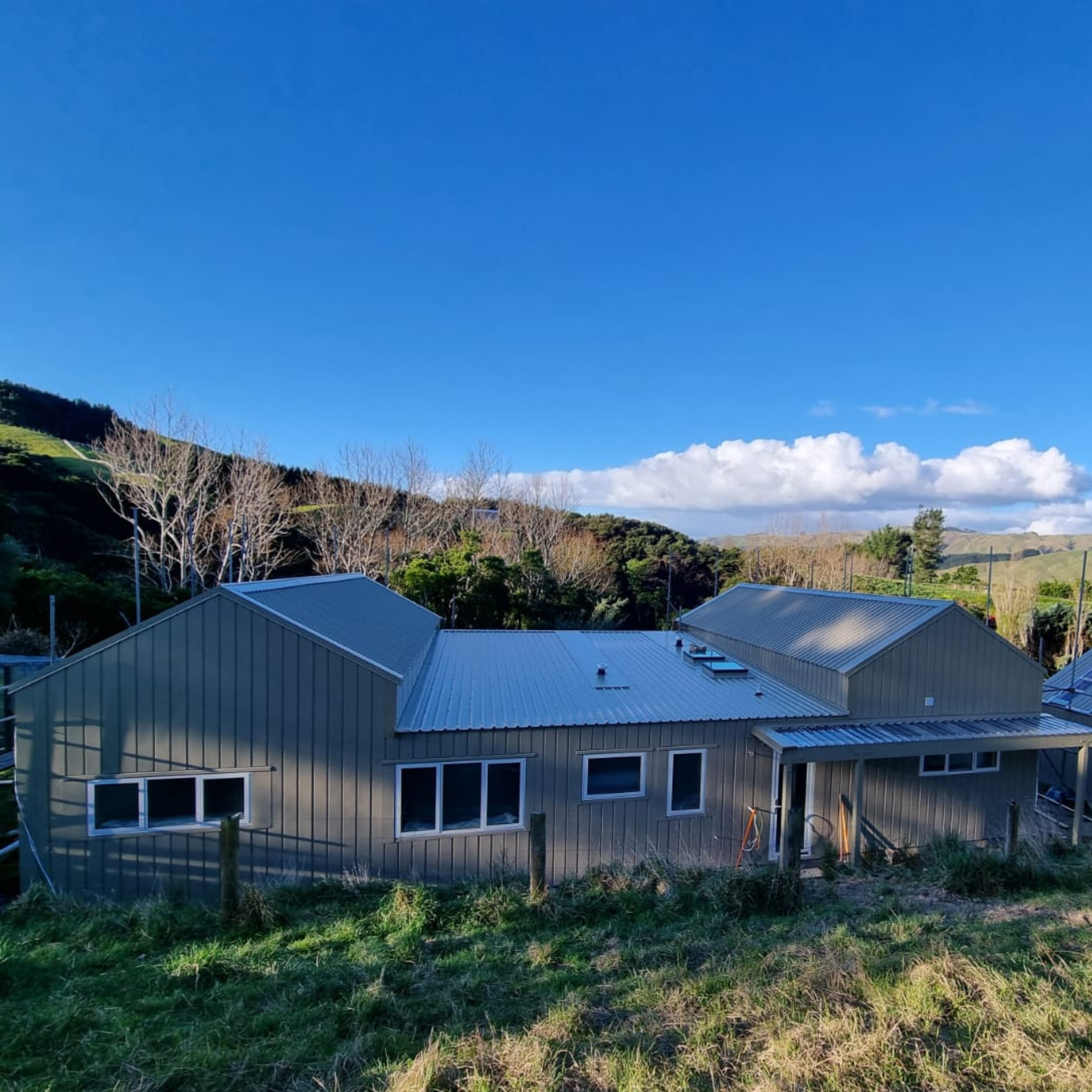 Residential buildings with metal roofs in a grassy area, under a clear blue sky with some clouds, surrounded by trees and hills in the background.