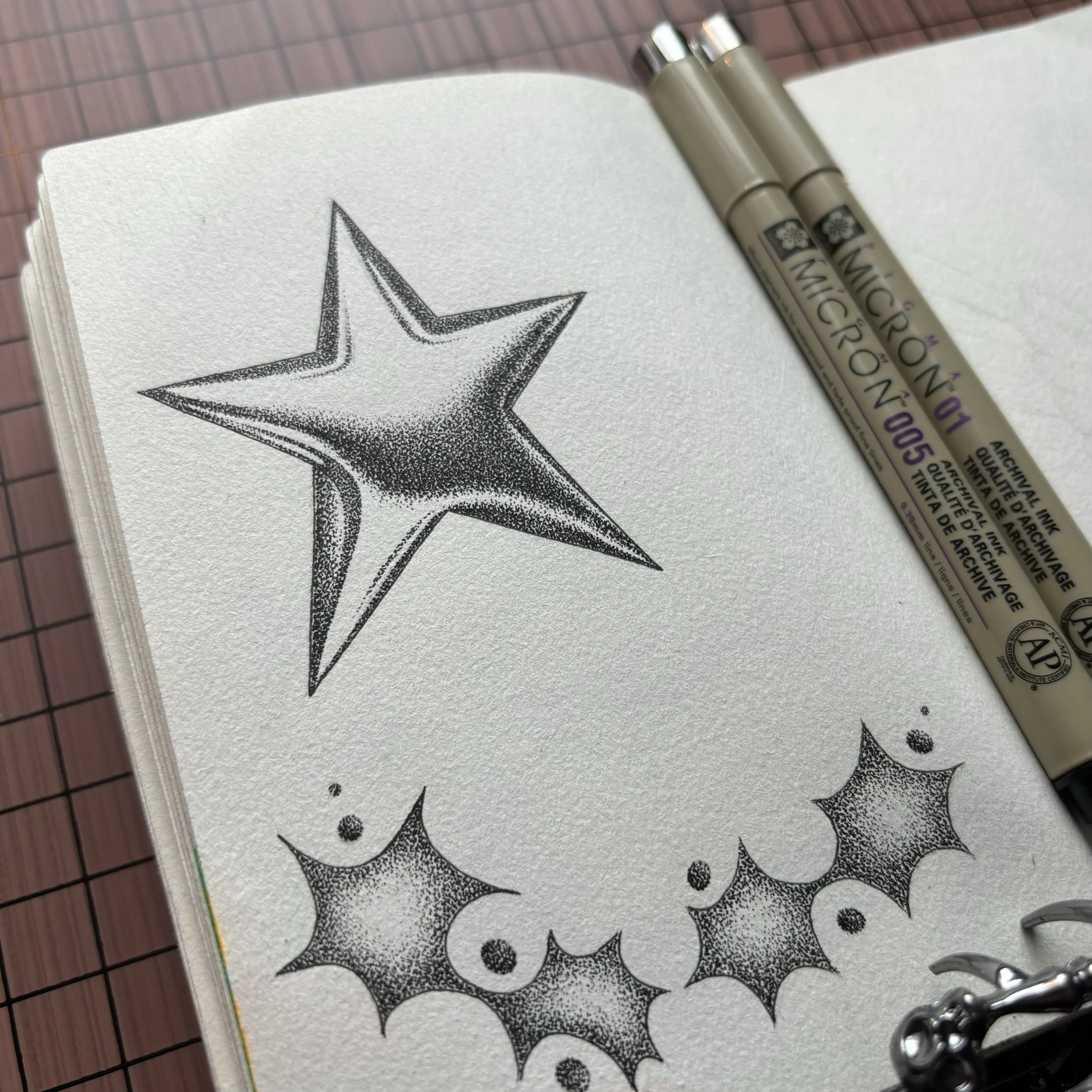 Black and white dot work style illustration of a star and holly leaves with berries, in a sketchbook with two beige ink pens nearby.