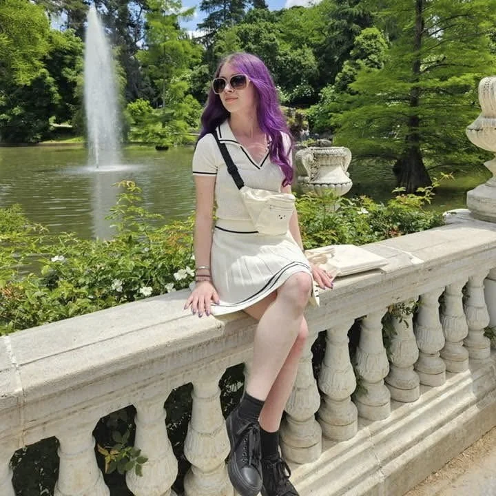 A young woman with purple hair, sunglasses, and a cream-colored outfit sitting on a stone railing in a park with a fountain and trees in the background.