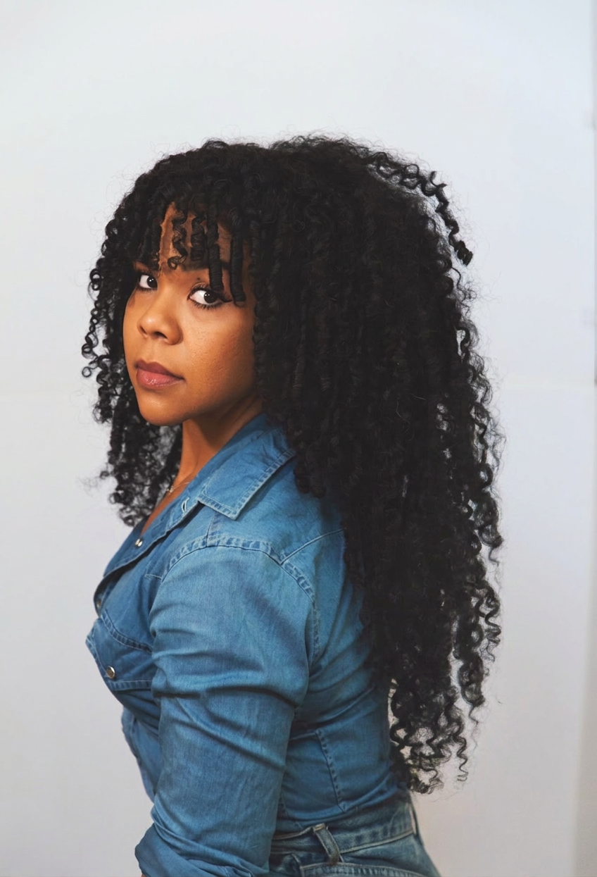 A woman with long, curly black hair wears a blue denim jacket and looks over her shoulder at the camera against a plain white background.