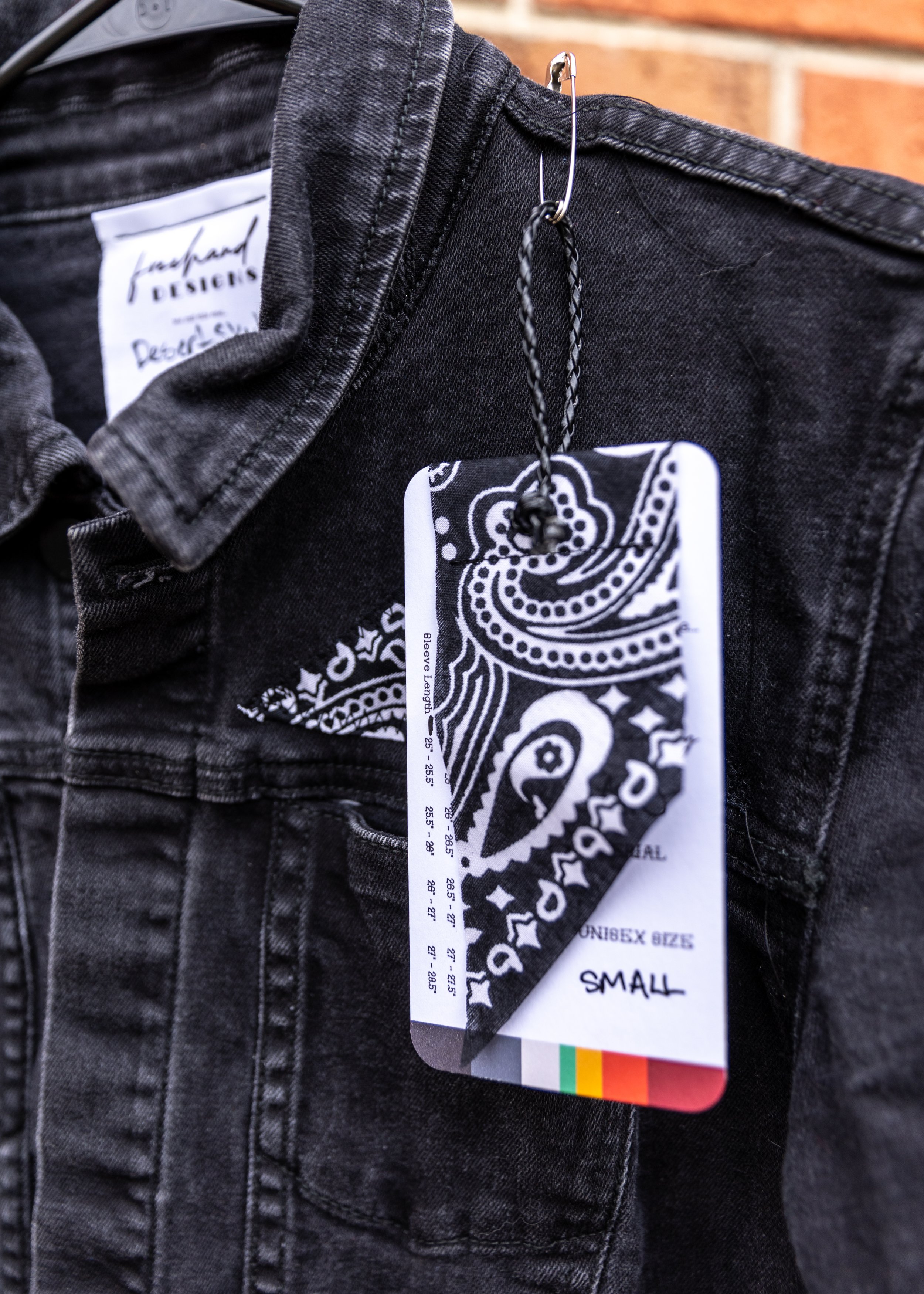 Close-up of black denim jeans with a striped black and white tag and two tags hanging from a safety pin, including a black and white bandana pattern and a size indicator reading 'small', against a brick wall background.