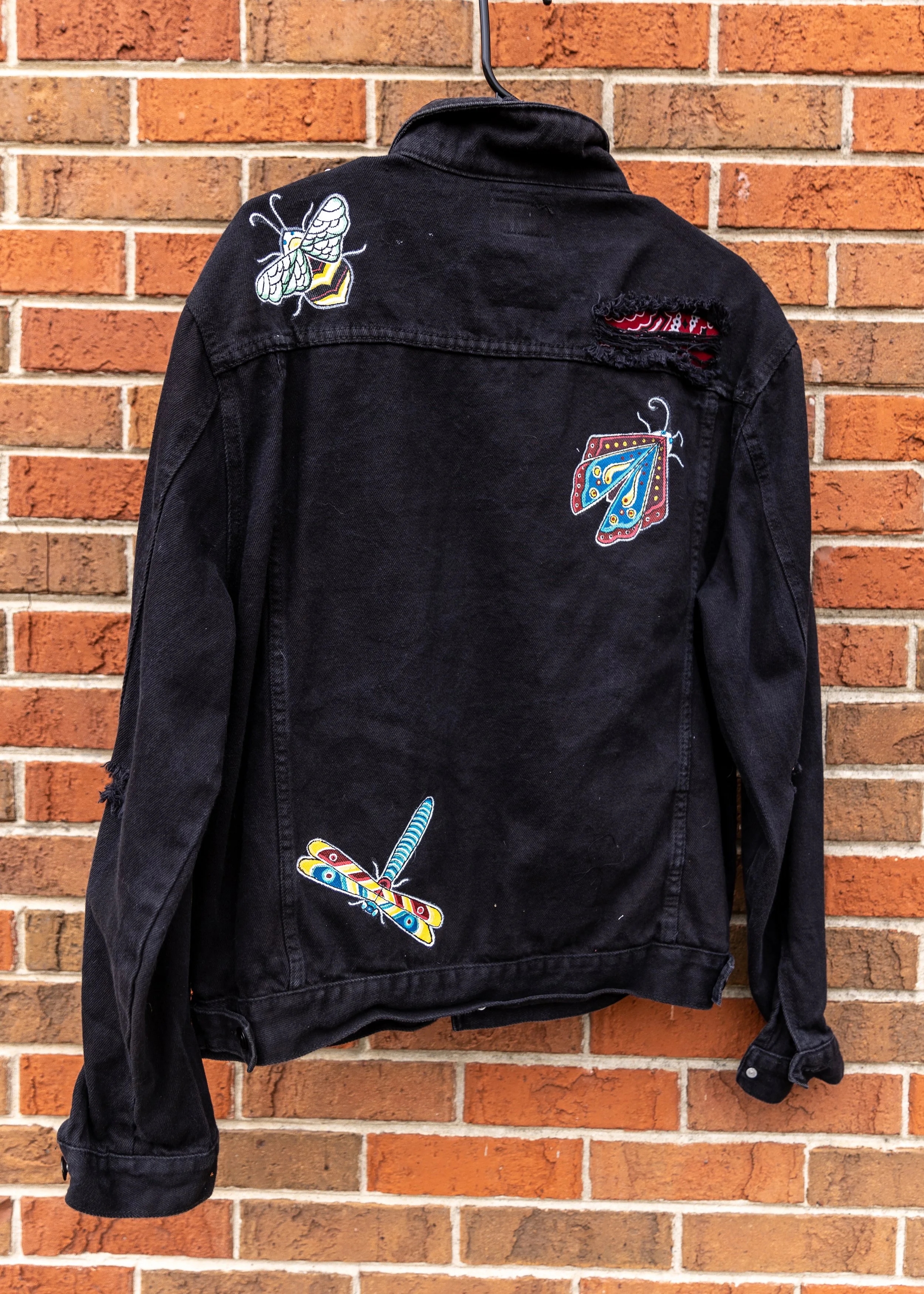 Black denim jacket with embroidered butterfly patches hanging against a red brick wall.