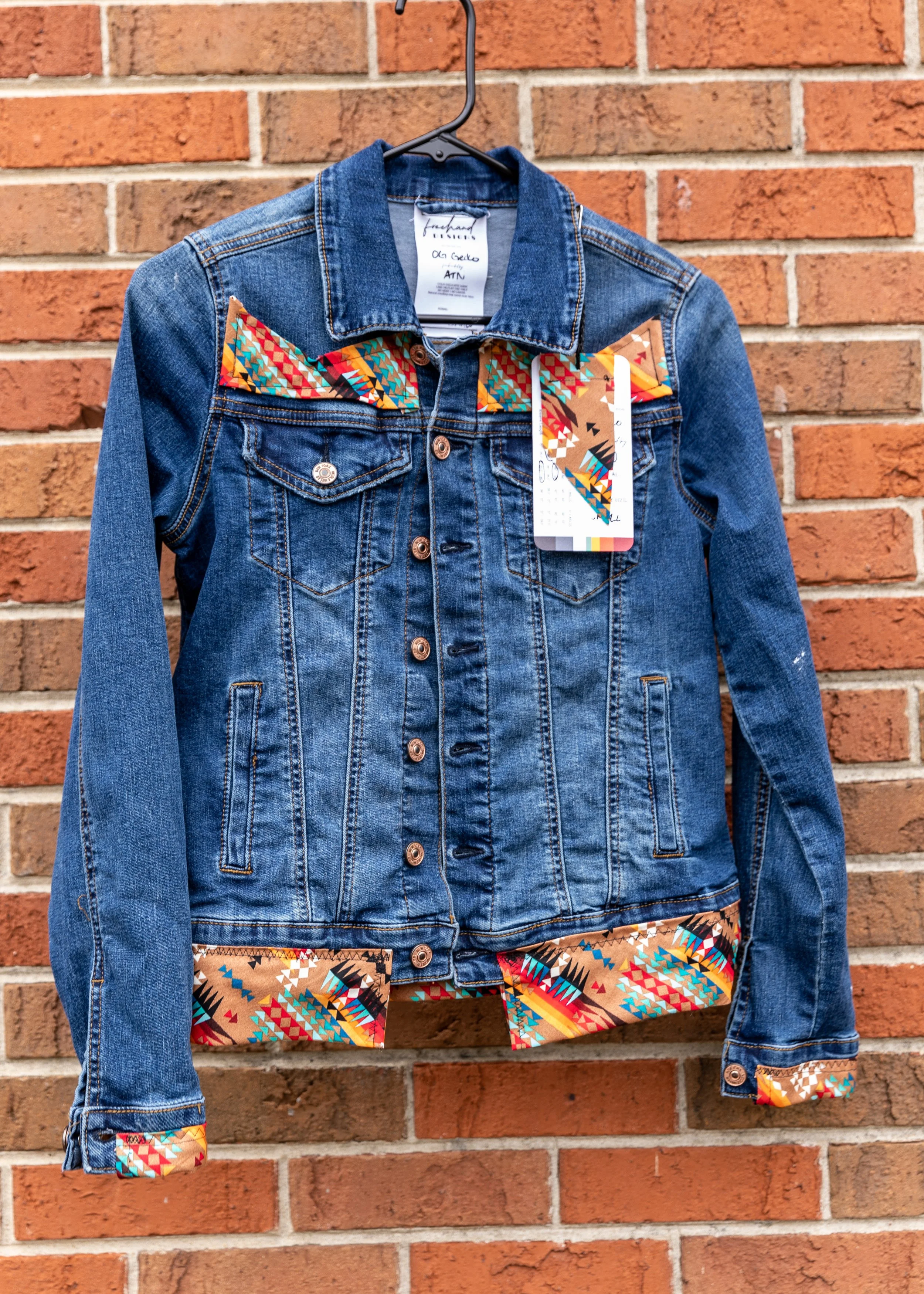 A denim jacket with colorful geometric patterned fabric accents on the collar, pockets, and cuffs, hanging on a black hanger against a red brick wall.