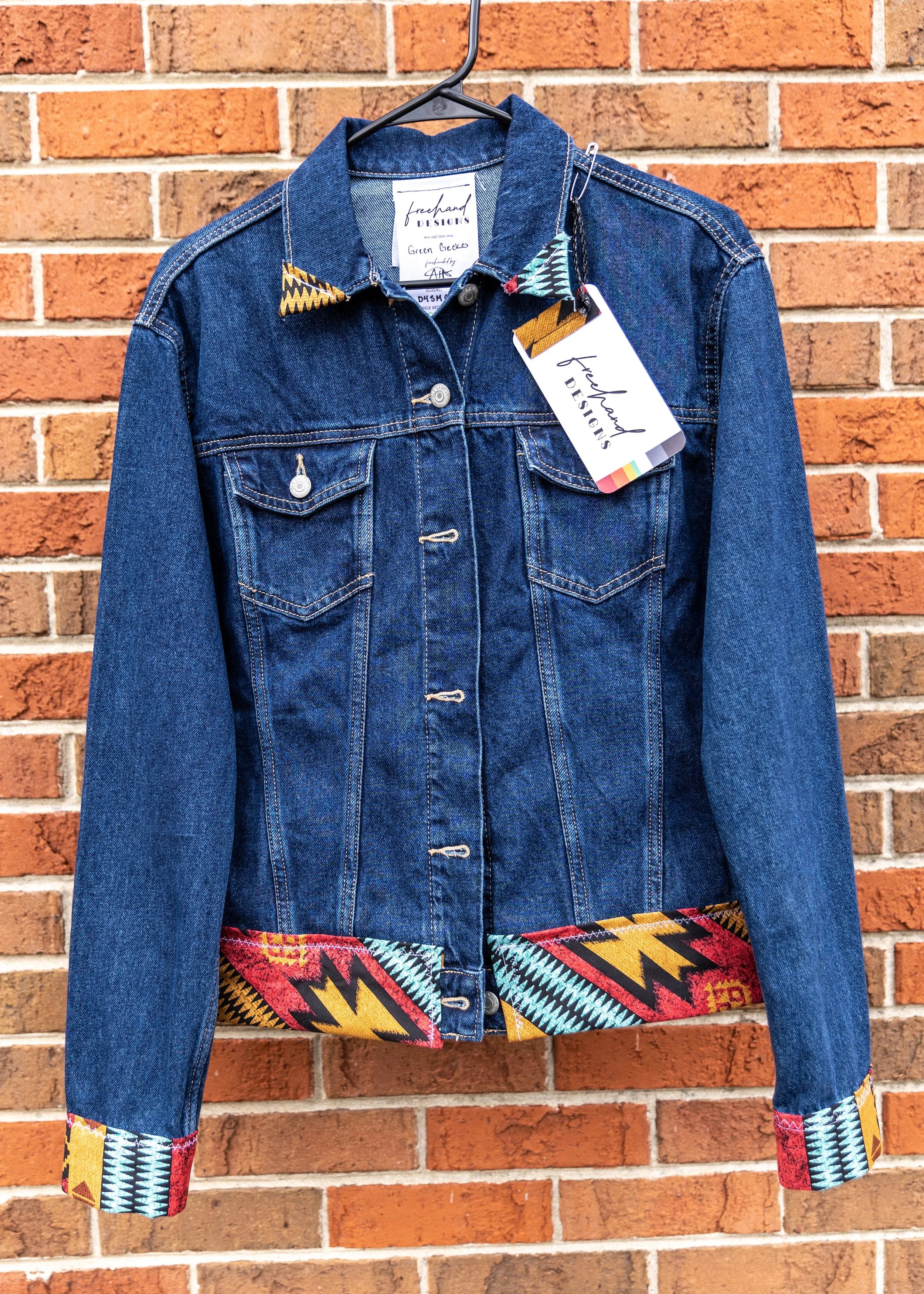 A denim jacket with colorful, patterned cuffs and collar hanging on a black hanger against a brick wall.