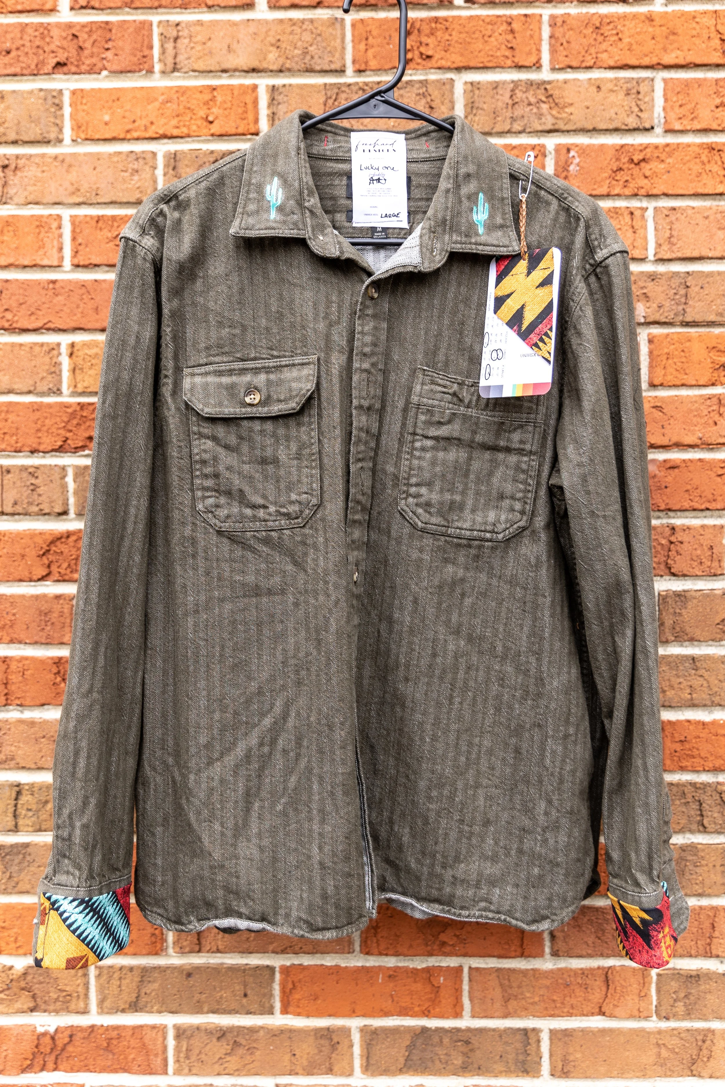 A long-sleeve, button-up shirt with two chest pockets, hanging on a black hanger against a brick wall. The shirt features a subtle vertical stripe pattern and has colorful, patterned fabric at the cuffs and collar with cactus embroidery.