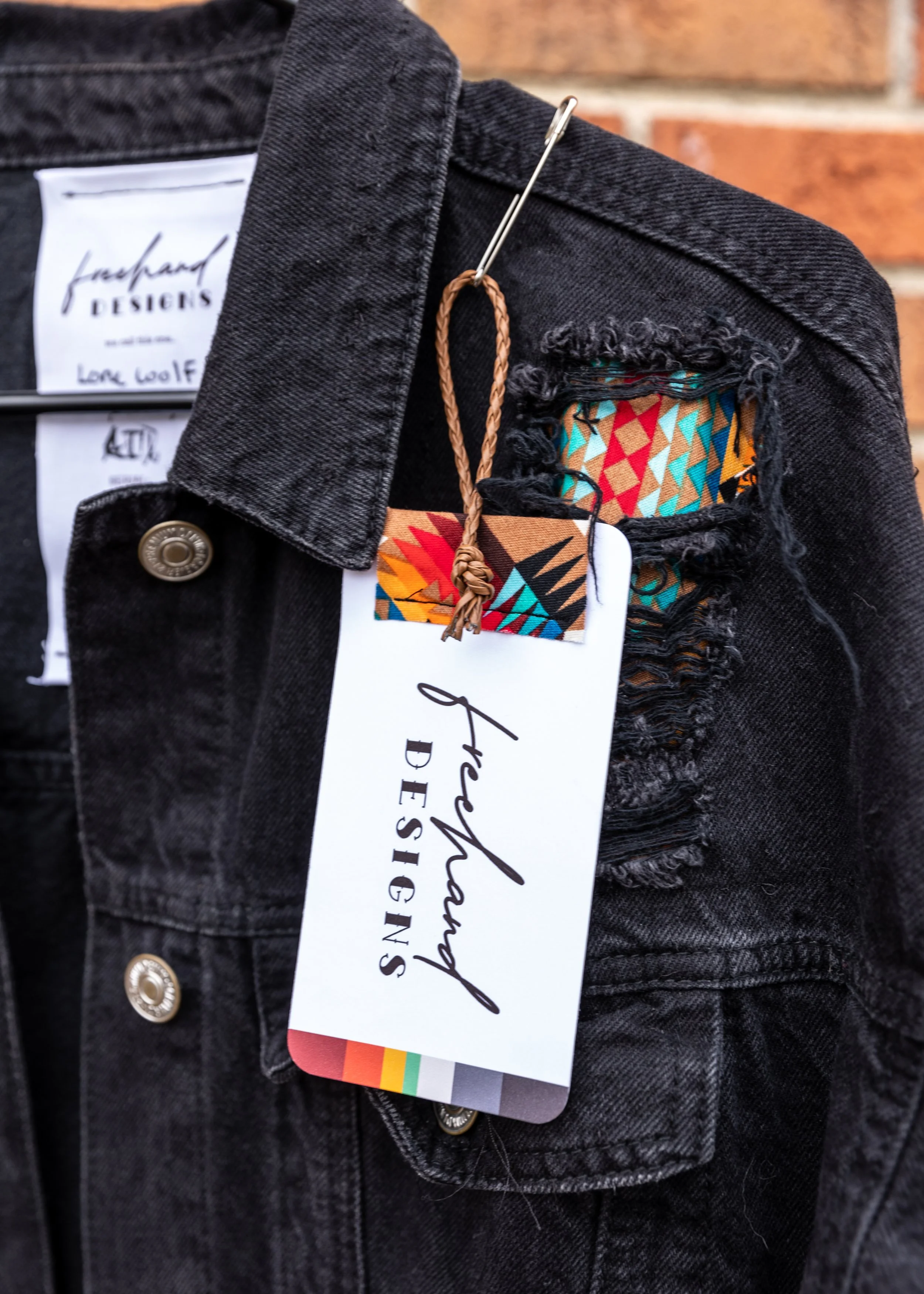 Black distressed denim jacket with a rainbow flag tag and a brown loop tag, hanging on a hook against a brick wall.