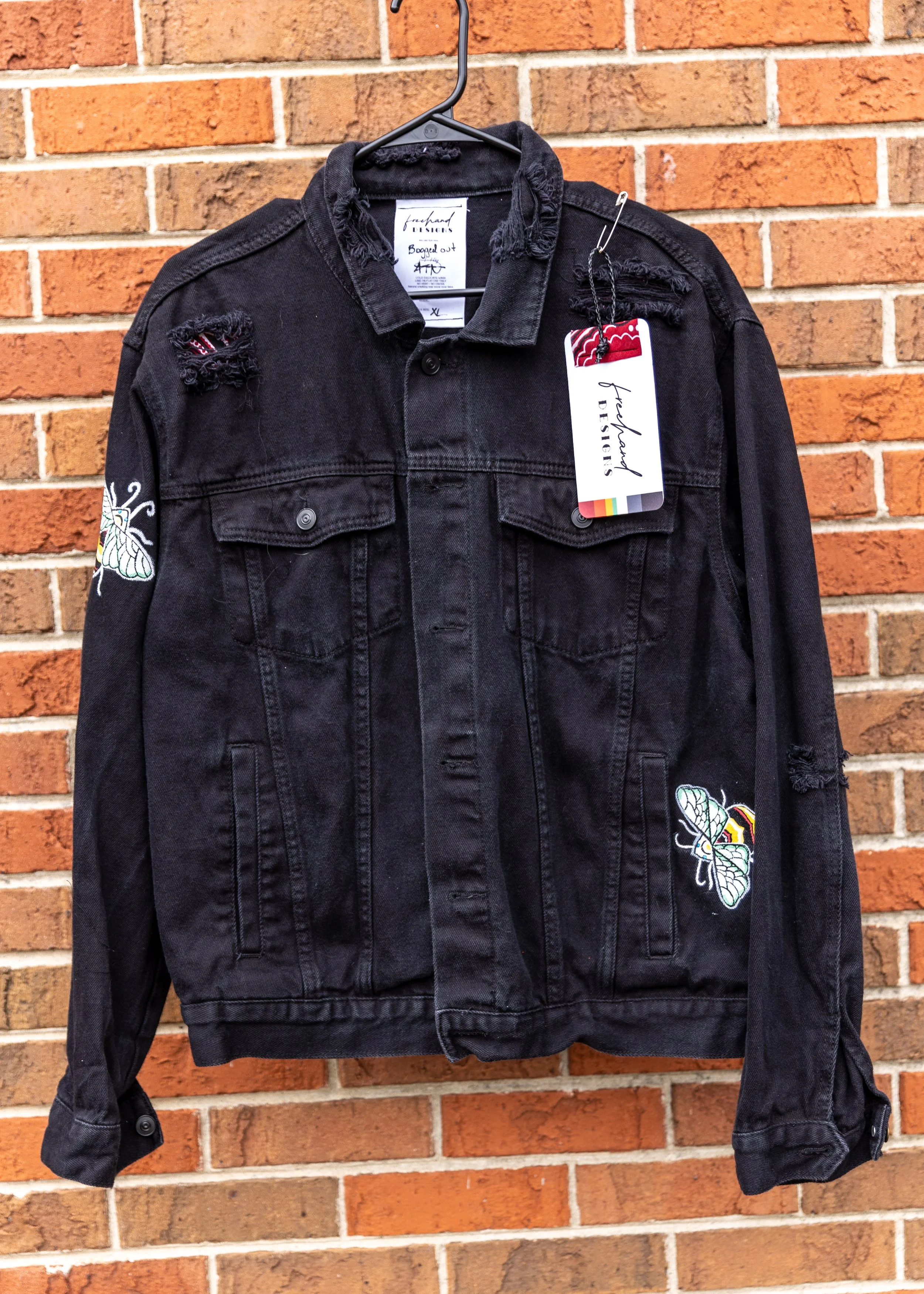 Black distressed denim jacket with butterfly patches hanging on a black hanger in front of a red brick wall.