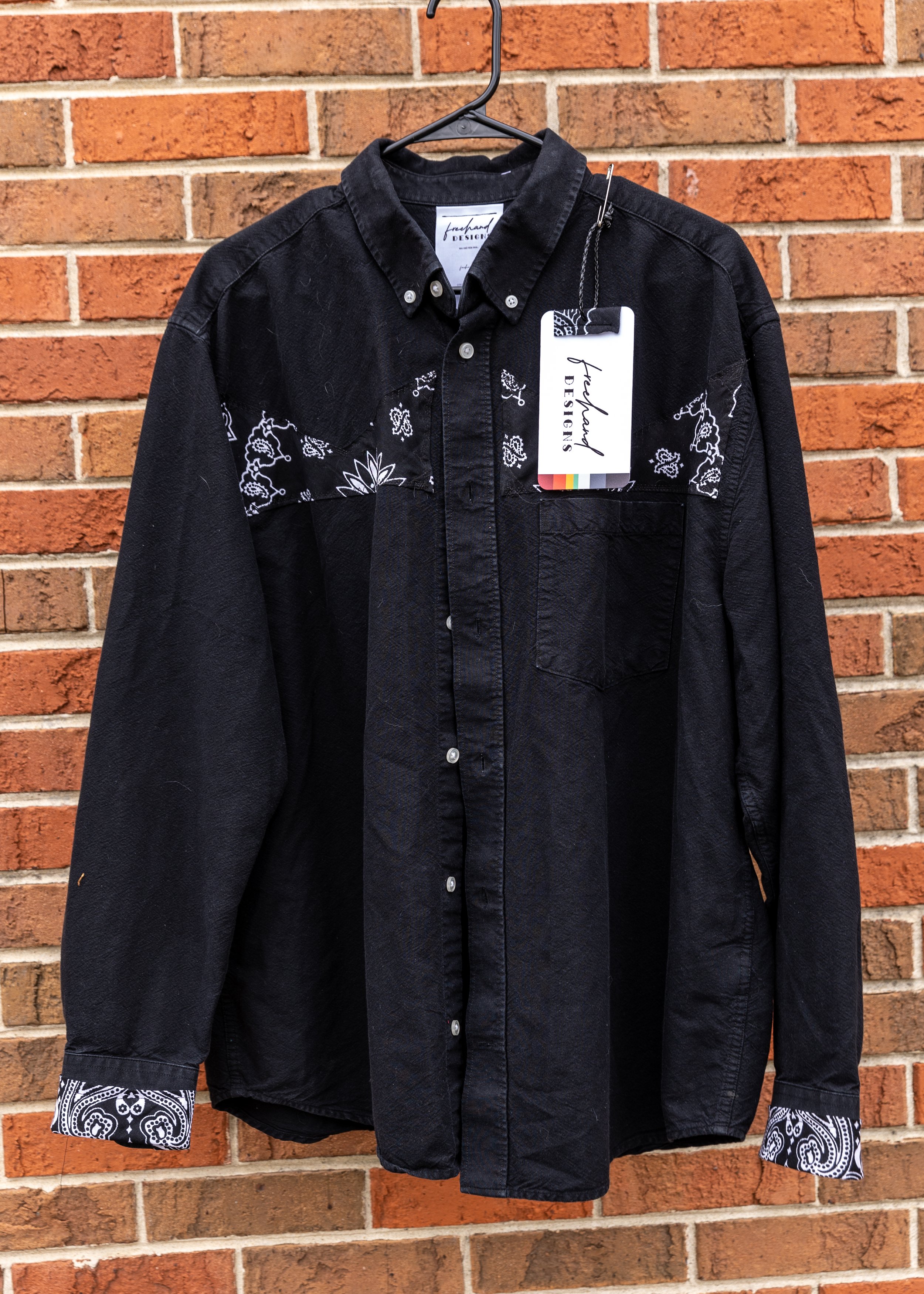Black button-down shirt with white paisley patterns on the collar and cuffs, hanging on a black hanger against a brick wall.