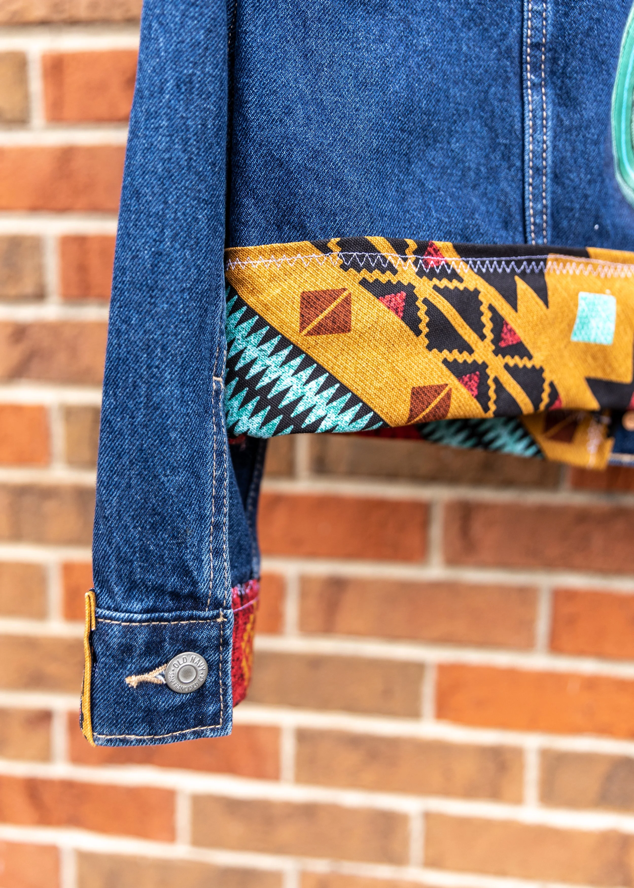Close-up of denim jacket sleeve, with colorful patterned fabric inside the sleeve, against a brick wall background.