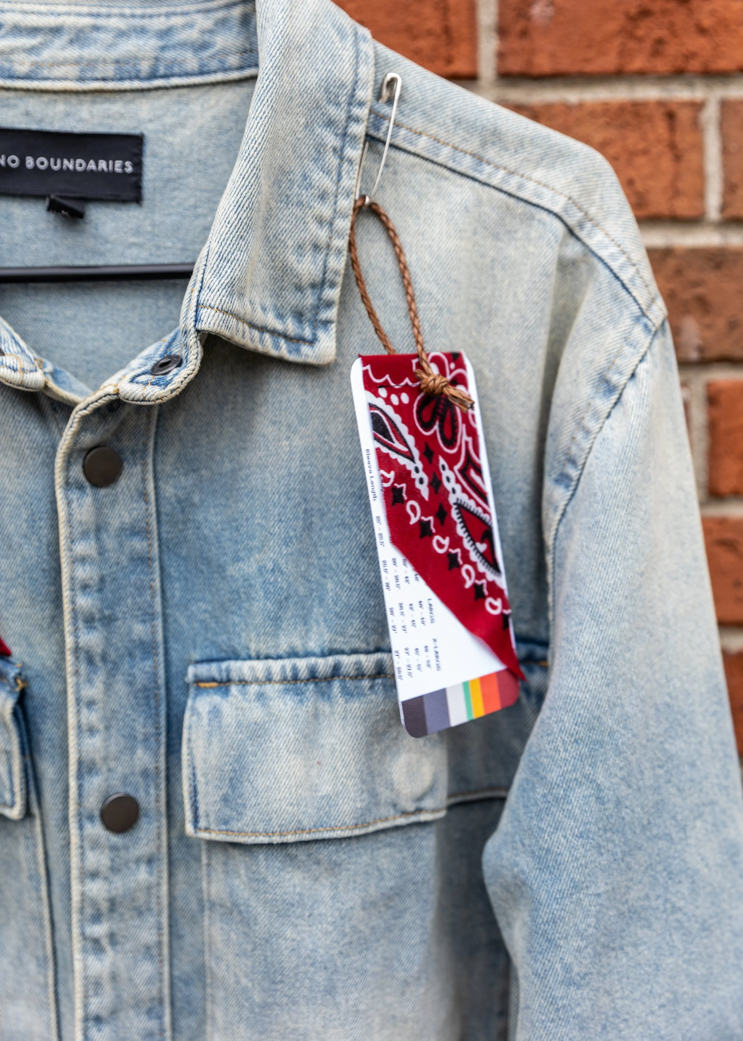 A close-up of a light blue denim jacket with a red, white, and black bandana hanging from a safety pin on the shoulder, with a brick wall in the background.