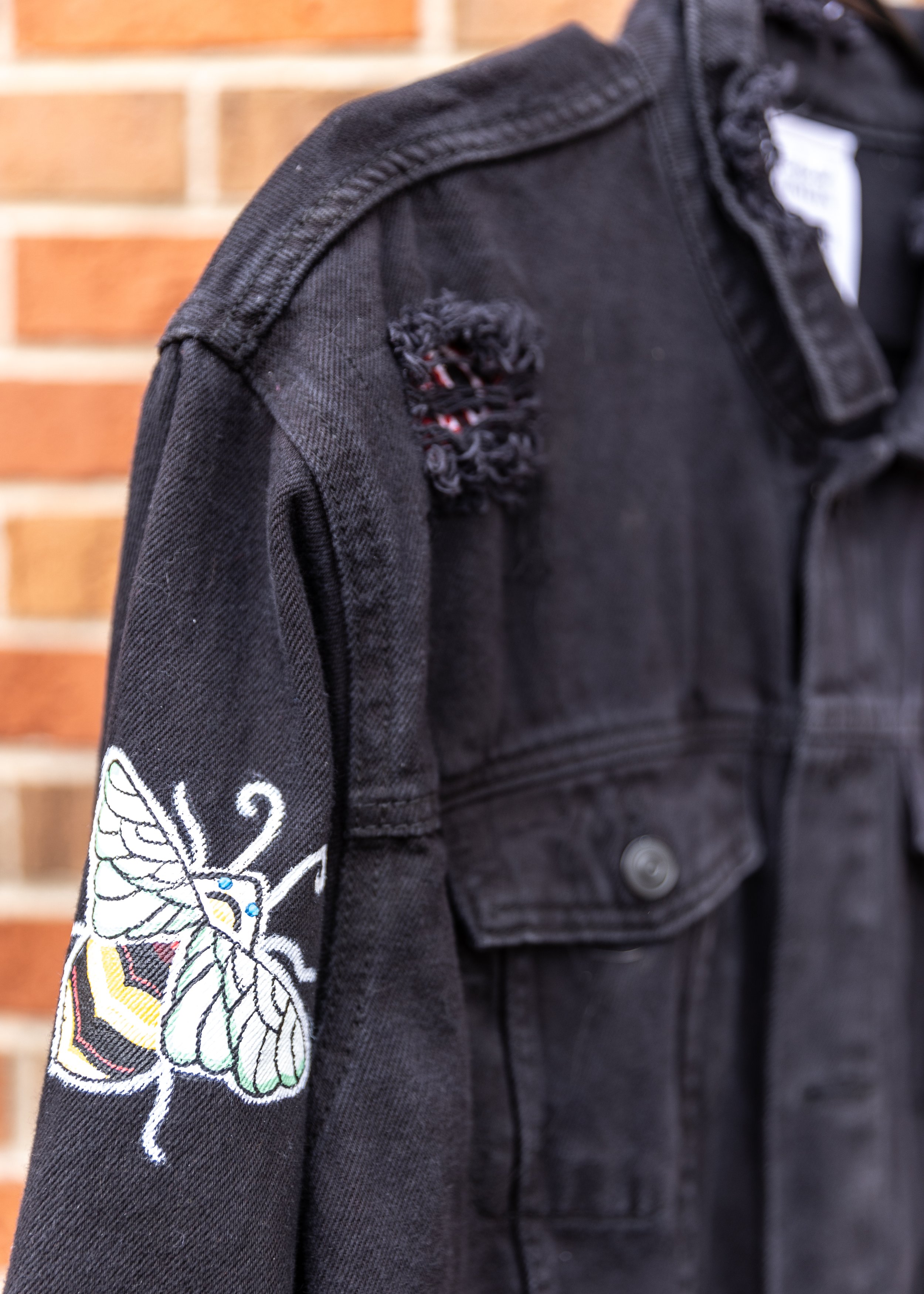 Black denim jacket with a colorful embroidered butterfly design on the sleeve, worn by a person standing in front of a brick wall.