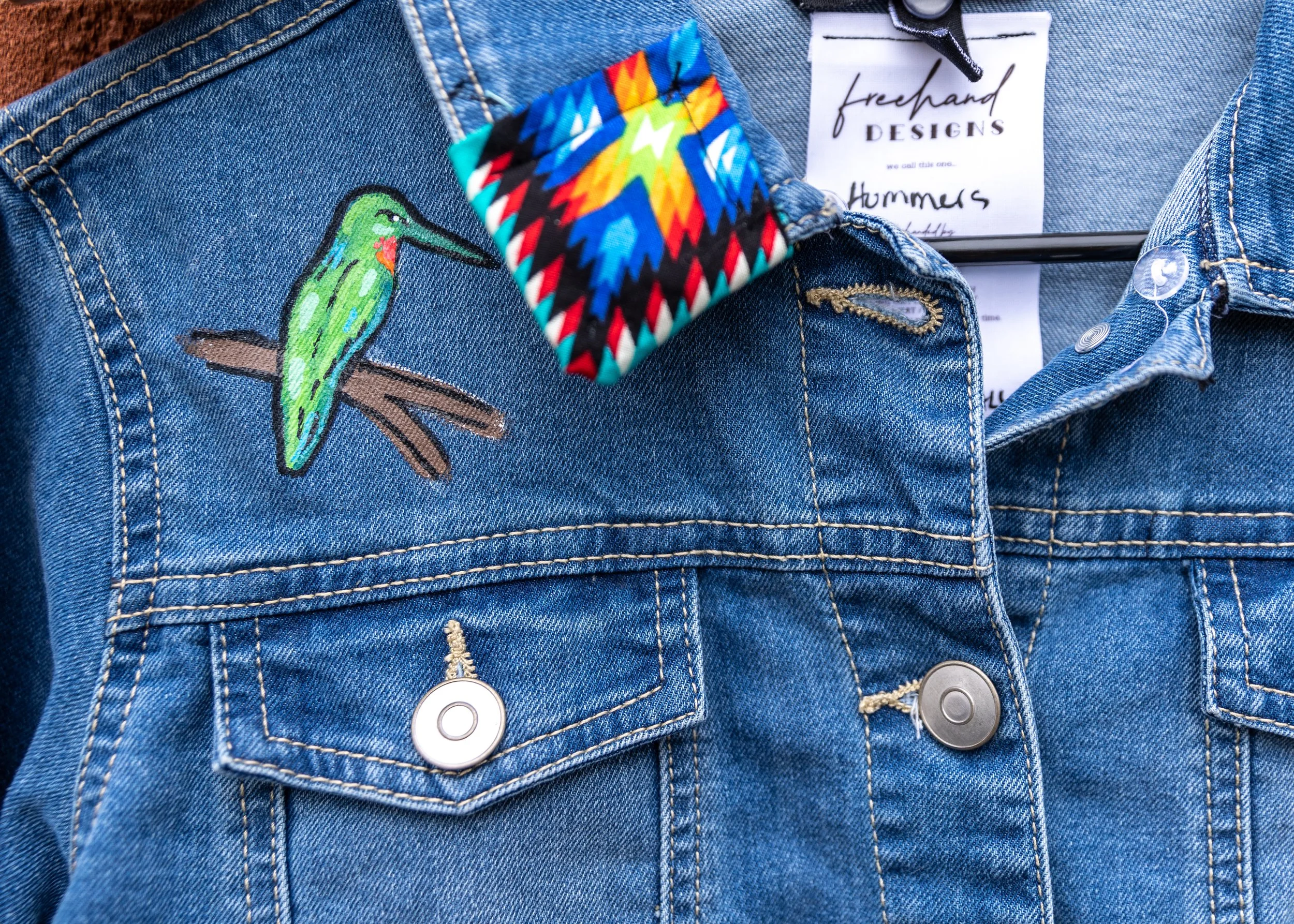 Close-up of a denim jacket with an embroidered green hummingbird patch on the left side and a colorful rainbow-patterned fabric cuff. The jacket features metal buttons and a white clothing label inside.