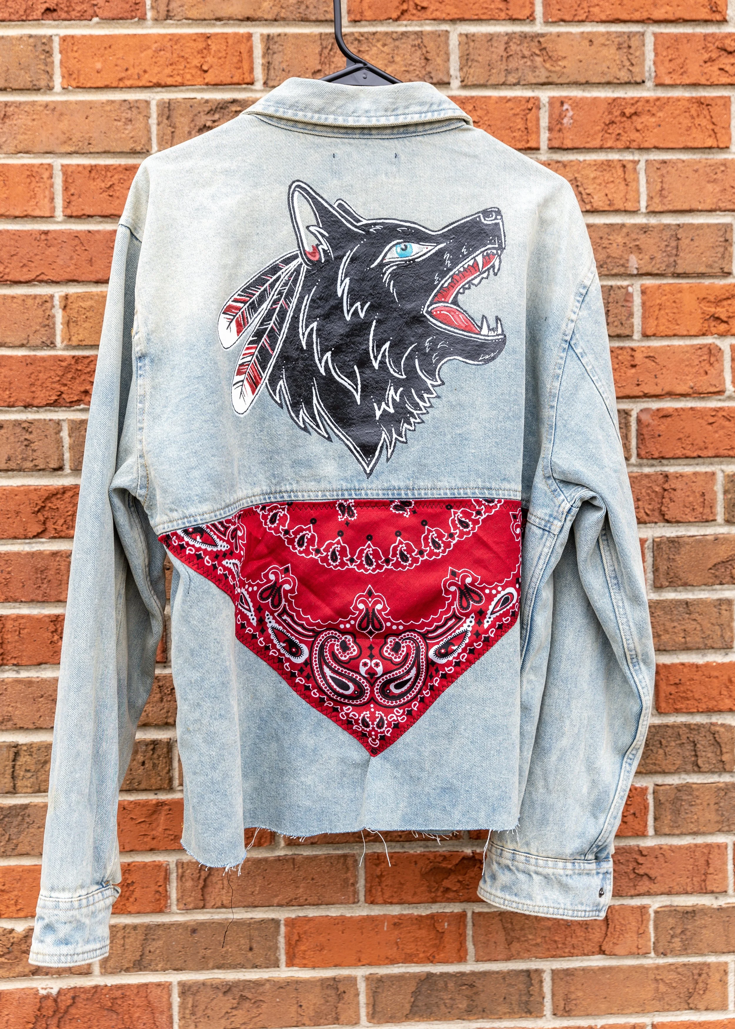 A denim jacket with a large embroidered design of a black wolf's head with blue eyes, red tongue, and feathers on the back, hanging on a black hanger against a brick wall, with a red bandana pattern sewn into the bottom.